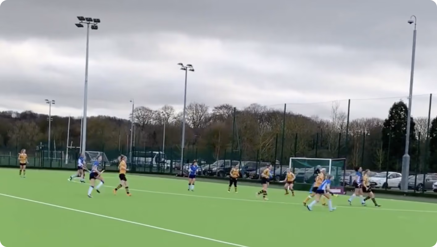 Field hockey players in yellow and blue uniforms competing on a green outdoor field under cloudy sky.
