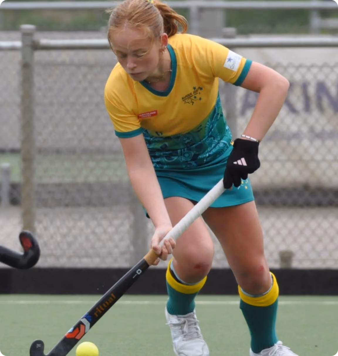 Female field hockey player in yellow and green uniform hitting a yellow ball with her stick.