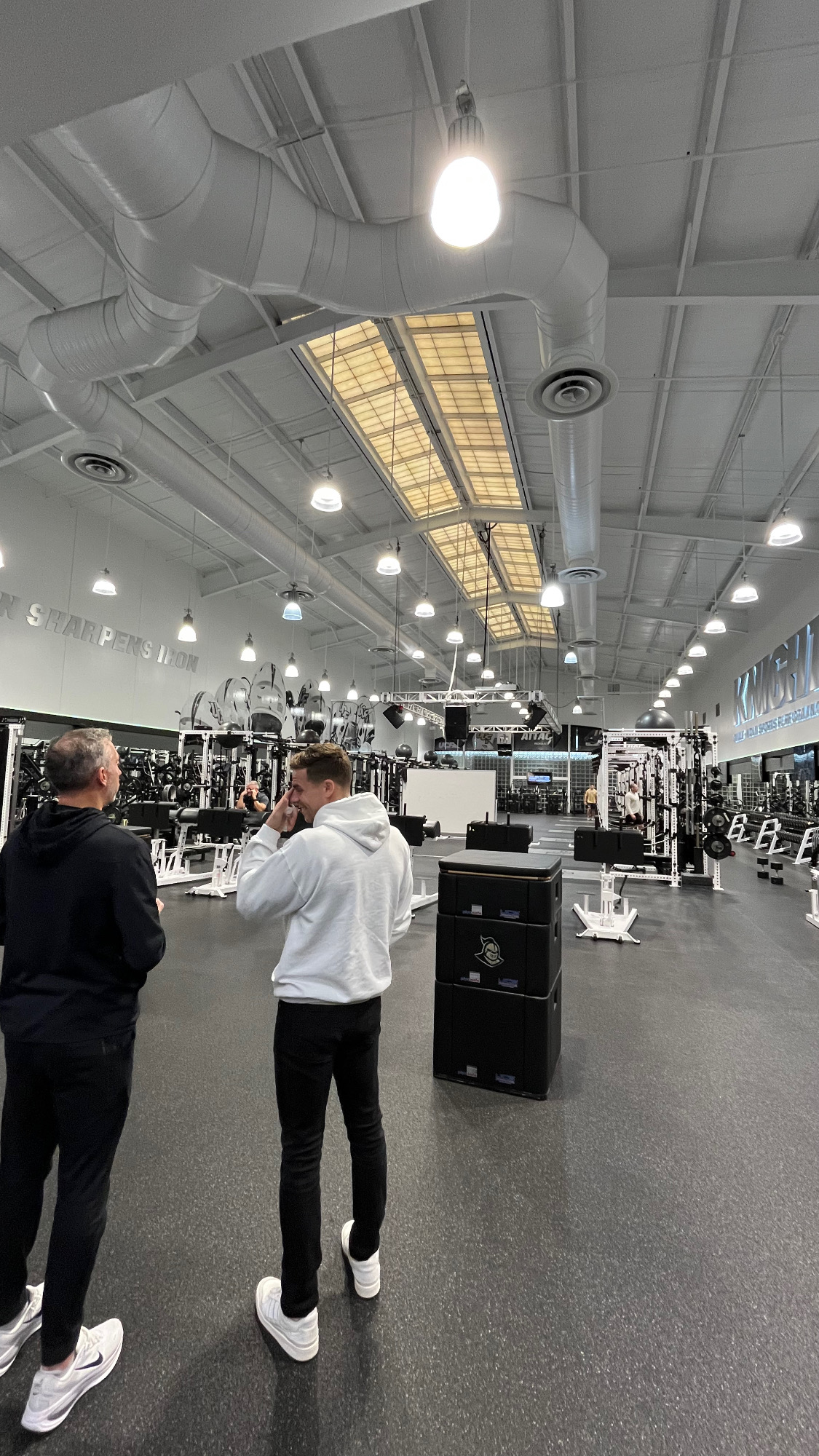 Two men standing inside a large gym with weightlifting equipment and high ceilings.