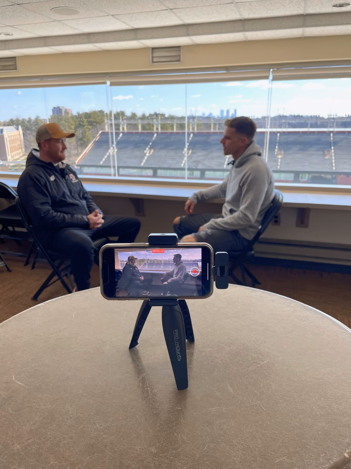 Two people having a conversation beside stadium windows while a phone on a tripod records.