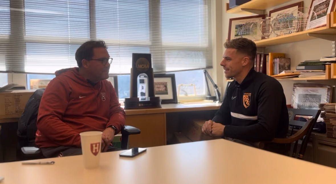 Two men seated across a desk in an office with books and sports memorabilia.