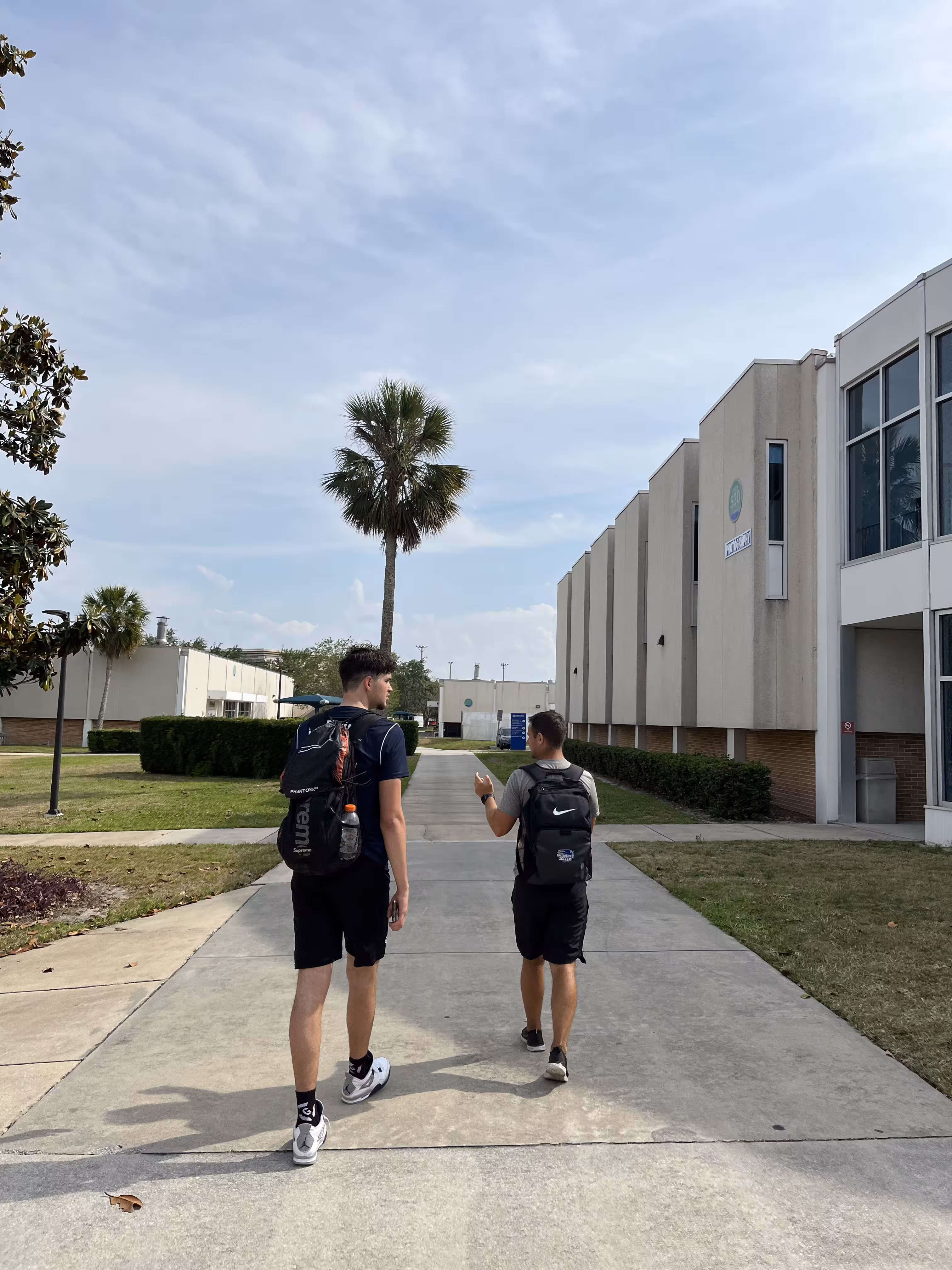 Two people carrying backpacks walk along a path beside buildings and palm trees.