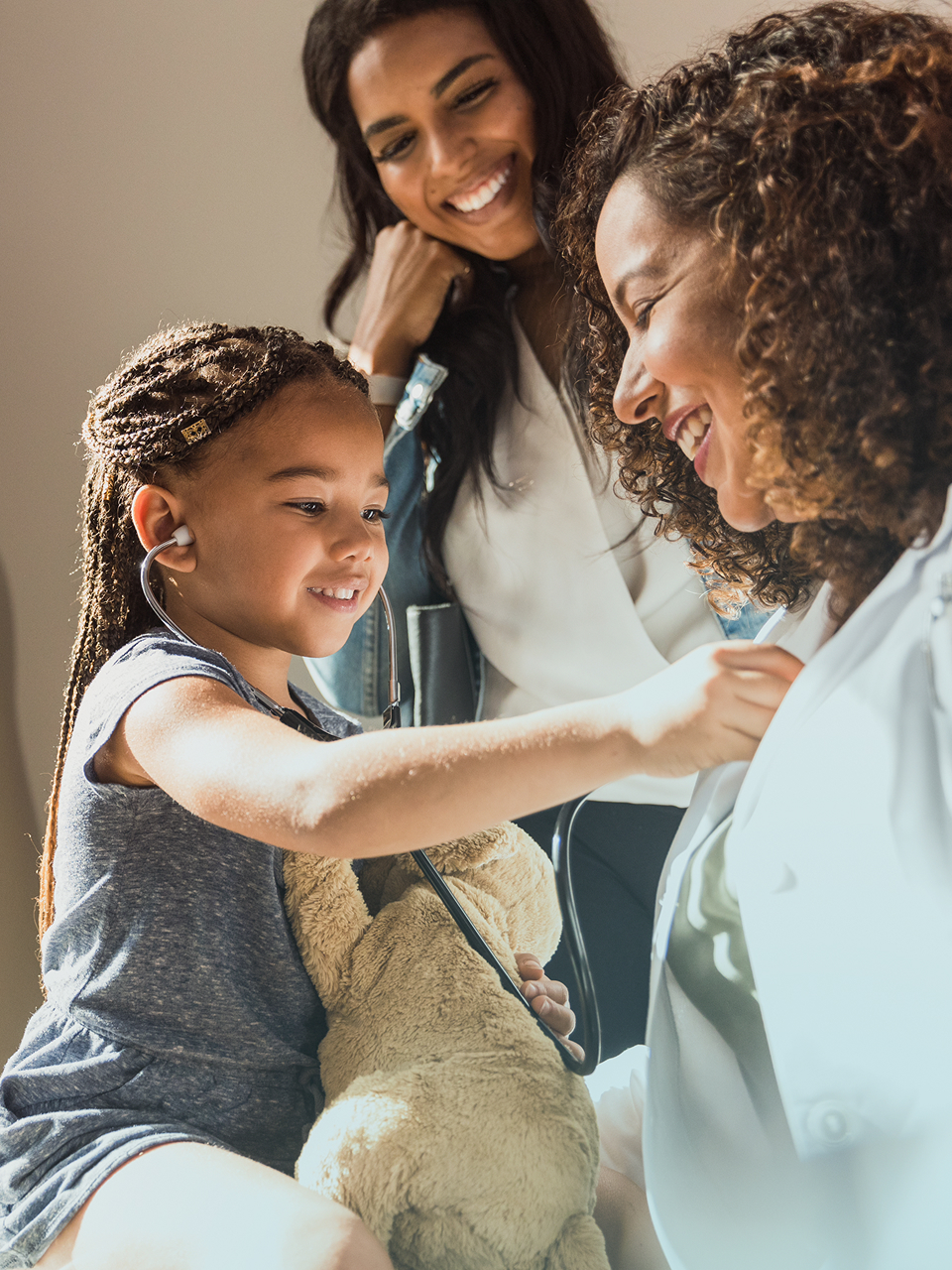 Smiling little girl uses a stethoscope on a female doctor while her mother watches happily.