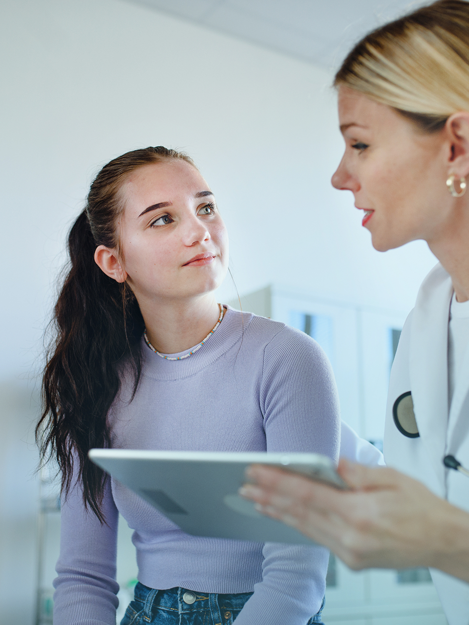 Young woman in lavender sweater listens attentively to a blonde healthcare professional holding a tablet.