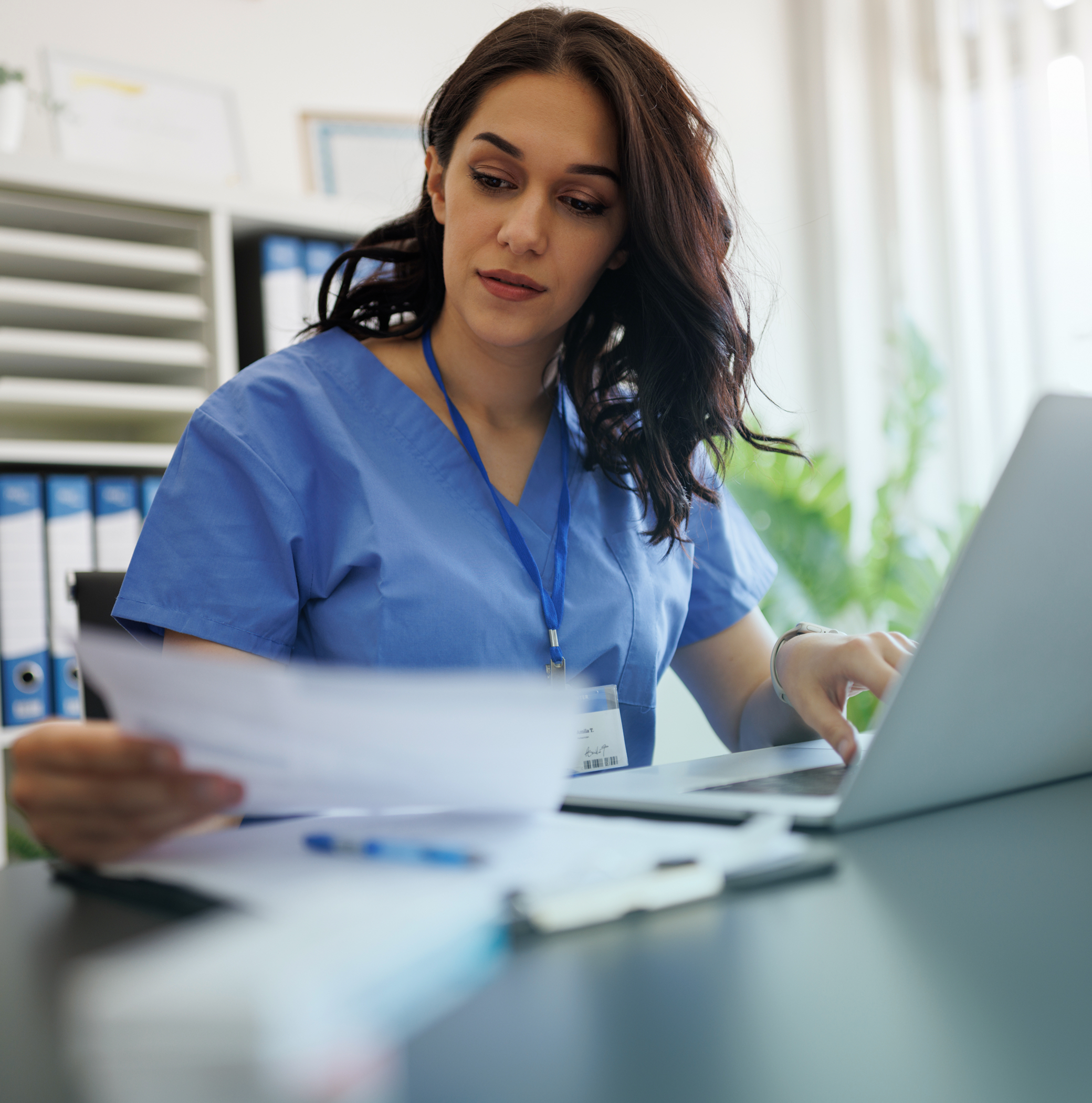 Healthcare worker in blue scrubs reviewing a document while using a laptop at a desk.
