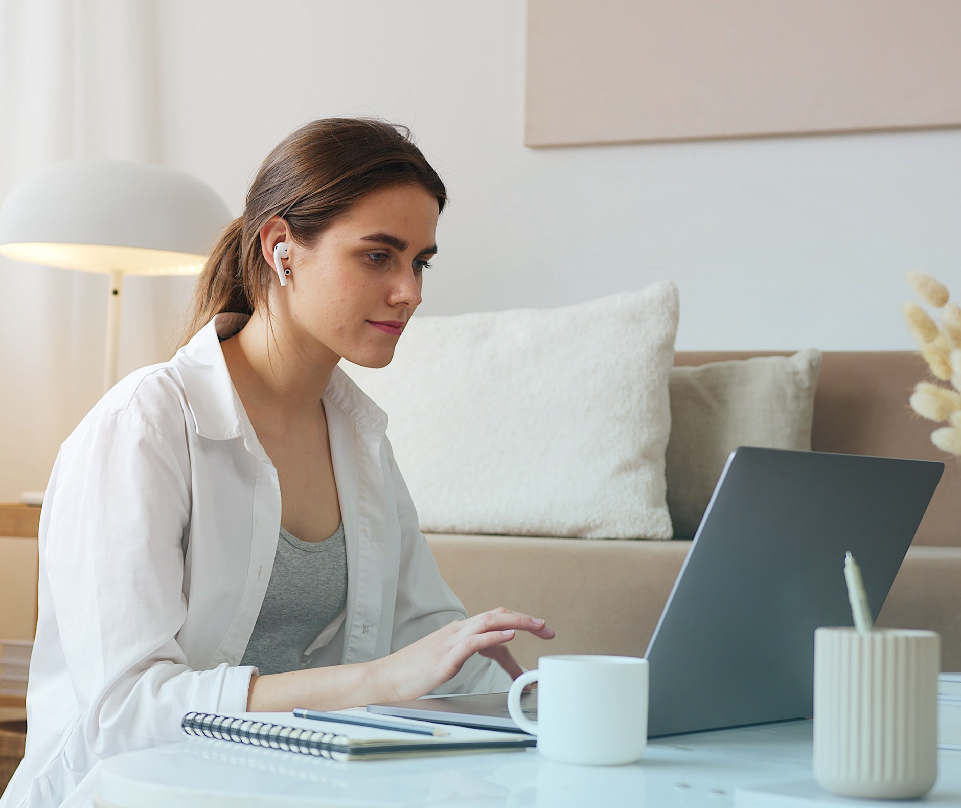 Woman wearing wireless earbuds working on a laptop at a table with a notebook and mug nearby.