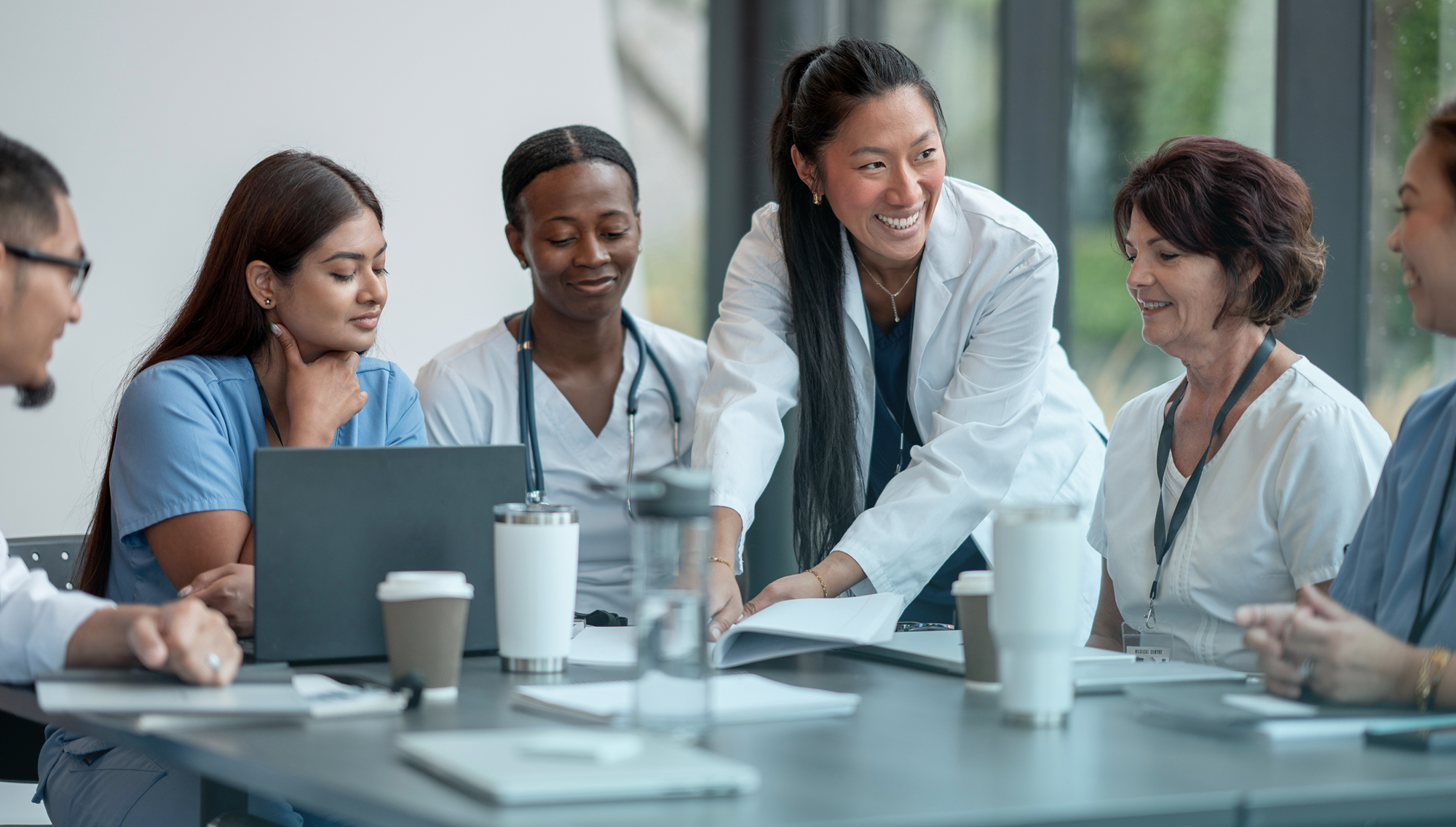 Group of healthcare professionals discussing around a table with laptop and papers in a conference room.