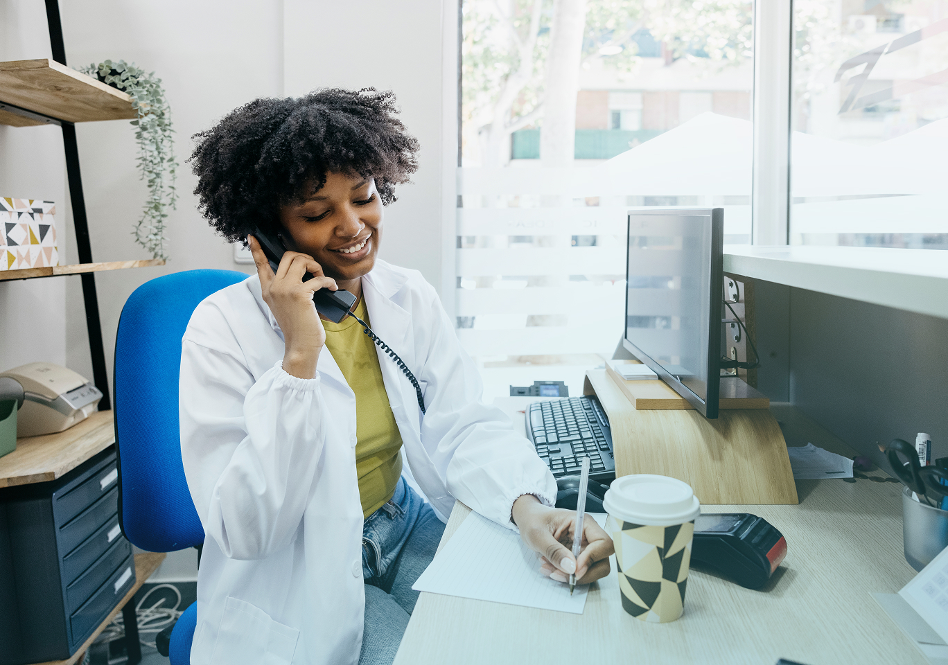 Smiling woman in white coat talking on phone and writing notes at desk with computer and coffee cup.