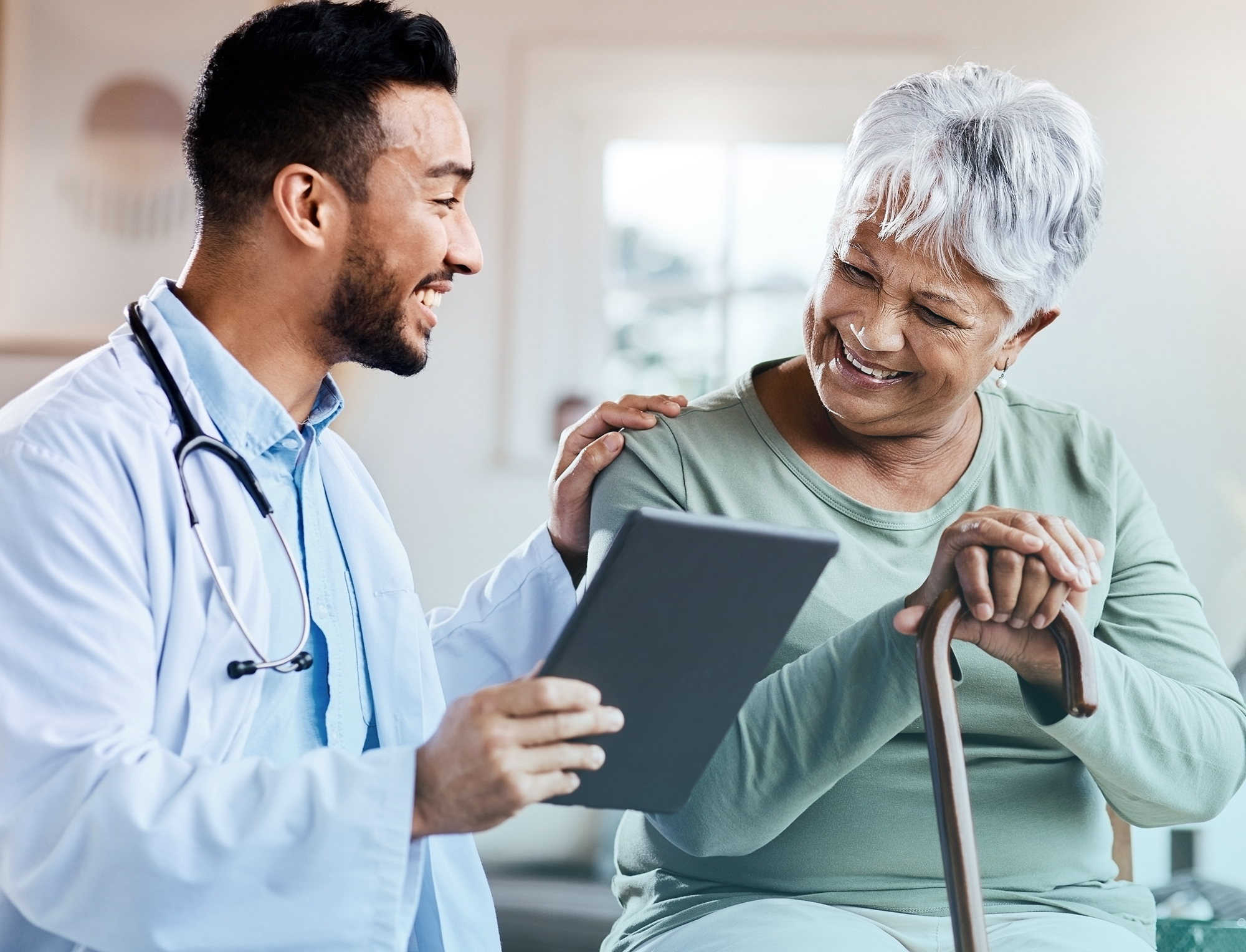 Smiling doctor holding tablet and comforting elderly woman with a cane in a bright room.