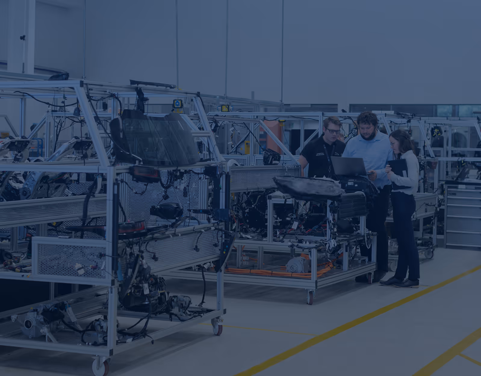 Three engineers reviewing data on a laptop in a factory with automotive parts and equipment on metal racks.