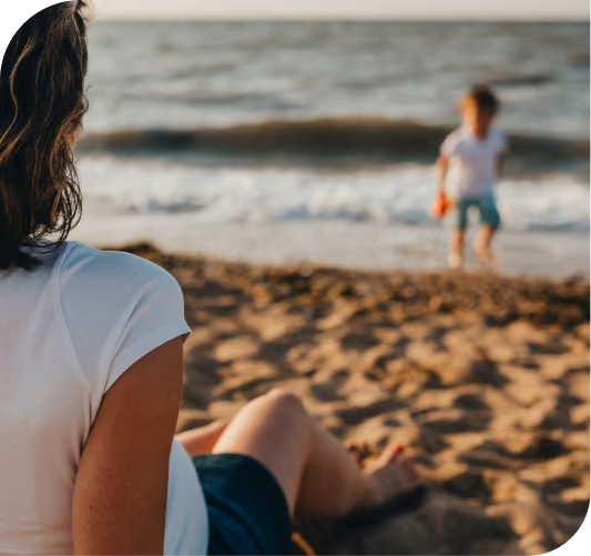 Woman sitting on sandy beach watching a child standing near the water's edge with waves in background.