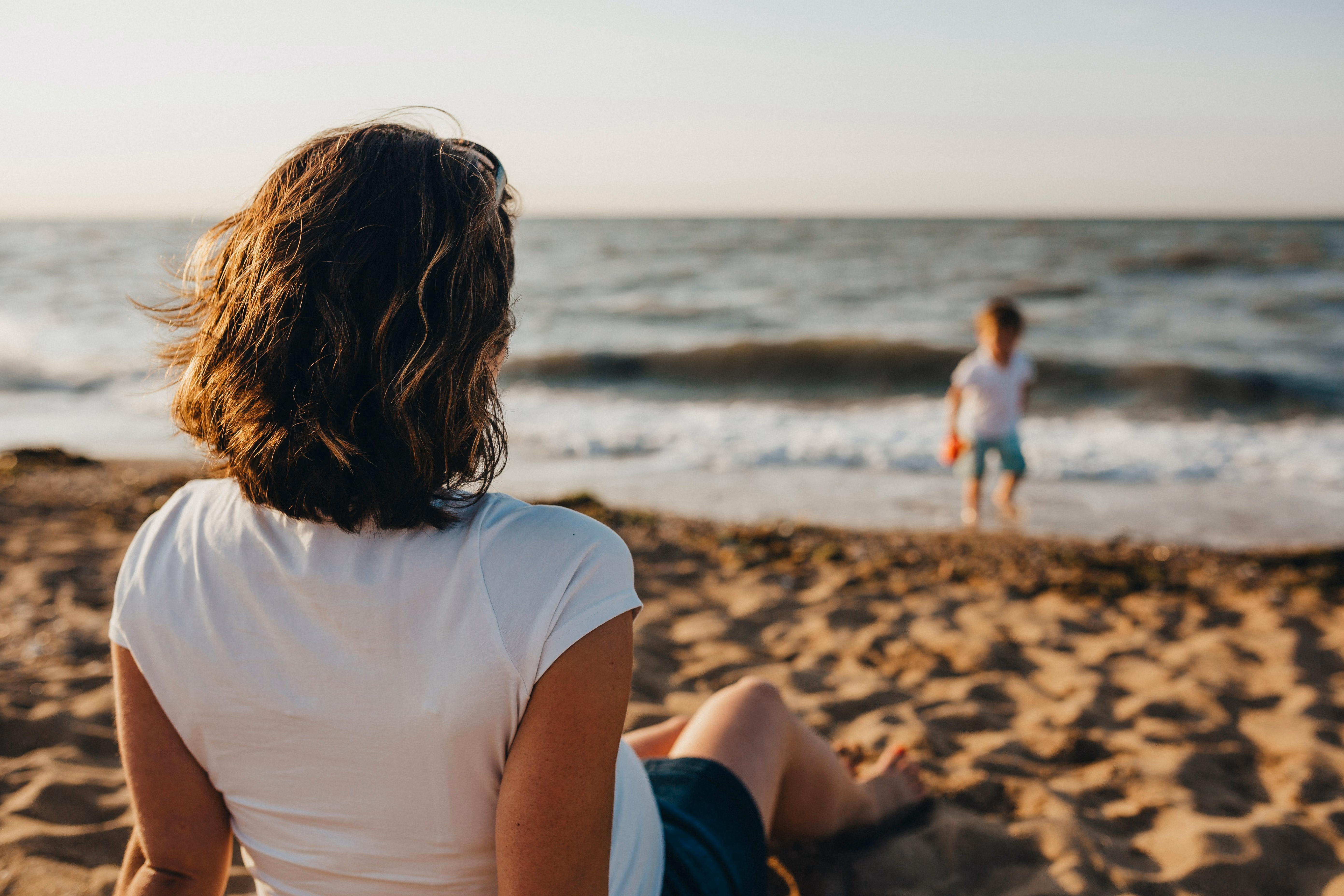 Woman sitting on sandy beach watching child playing near waves on a sunny day.