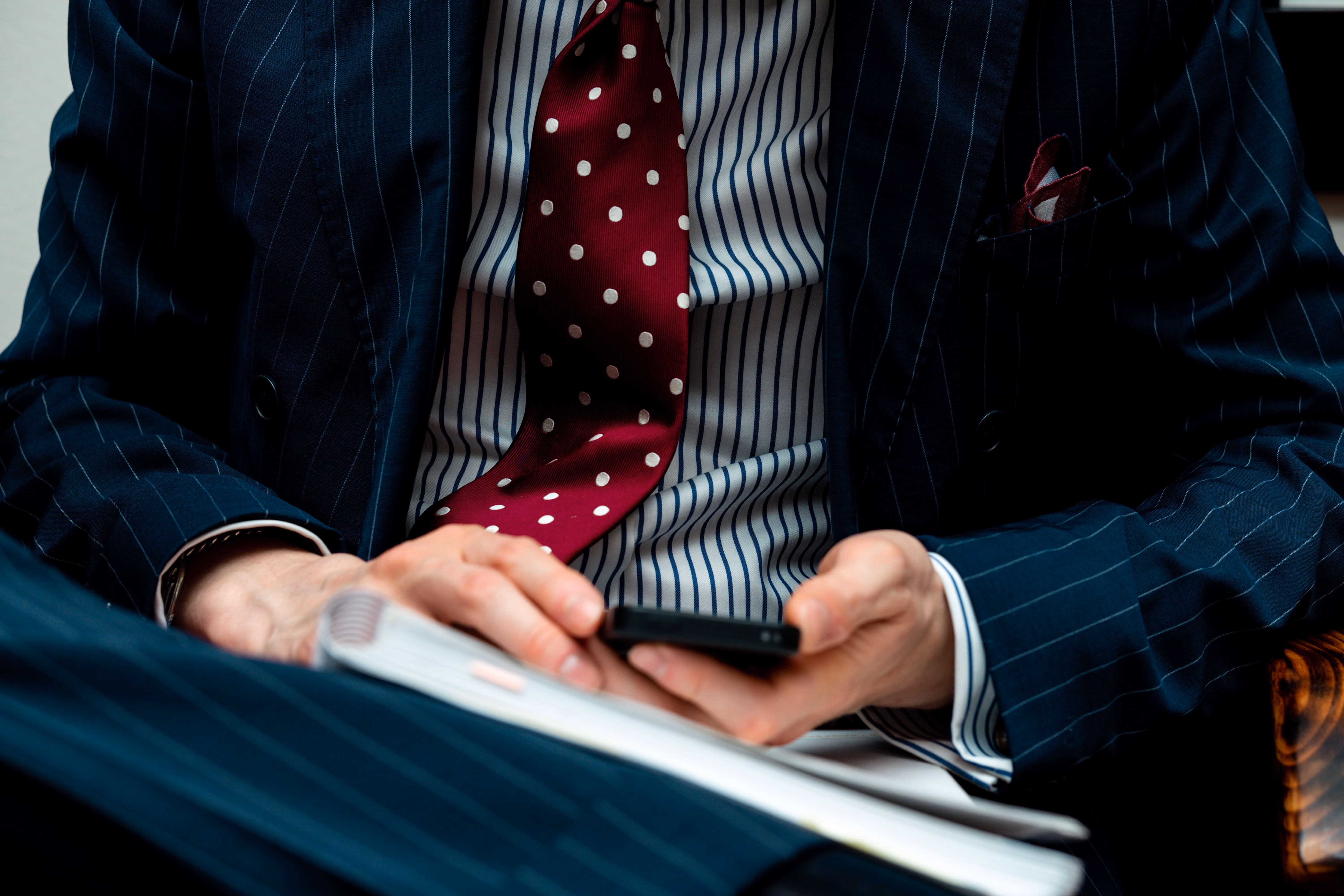 Close-up of person in pinstripe suit and polka dot tie holding a phone and notebook.