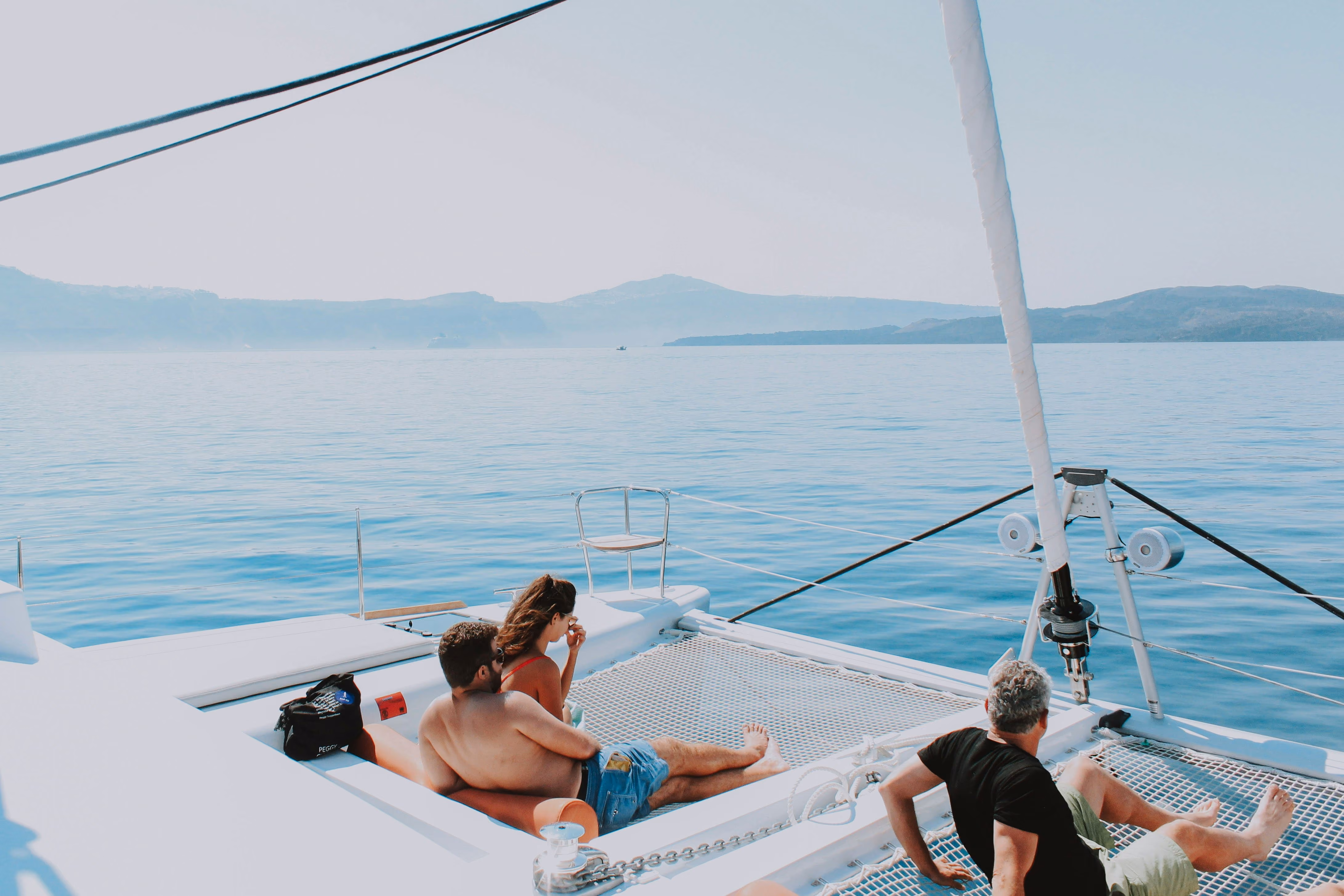 Three people relaxing on a white yacht netting with calm blue sea and distant hills under a clear sky.
