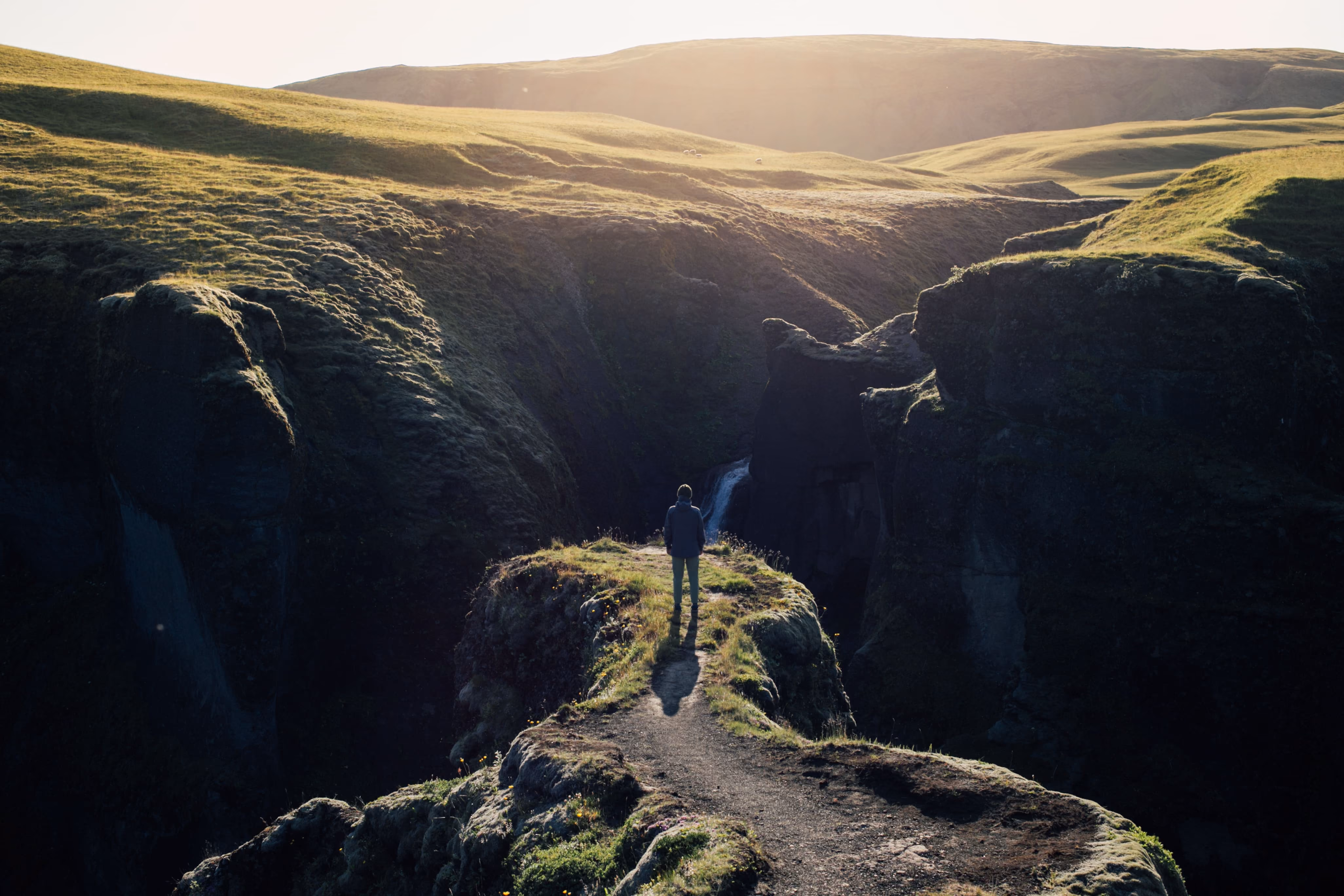 A person standing on a narrow grassy path overlooking cliffs with a waterfall in the background.