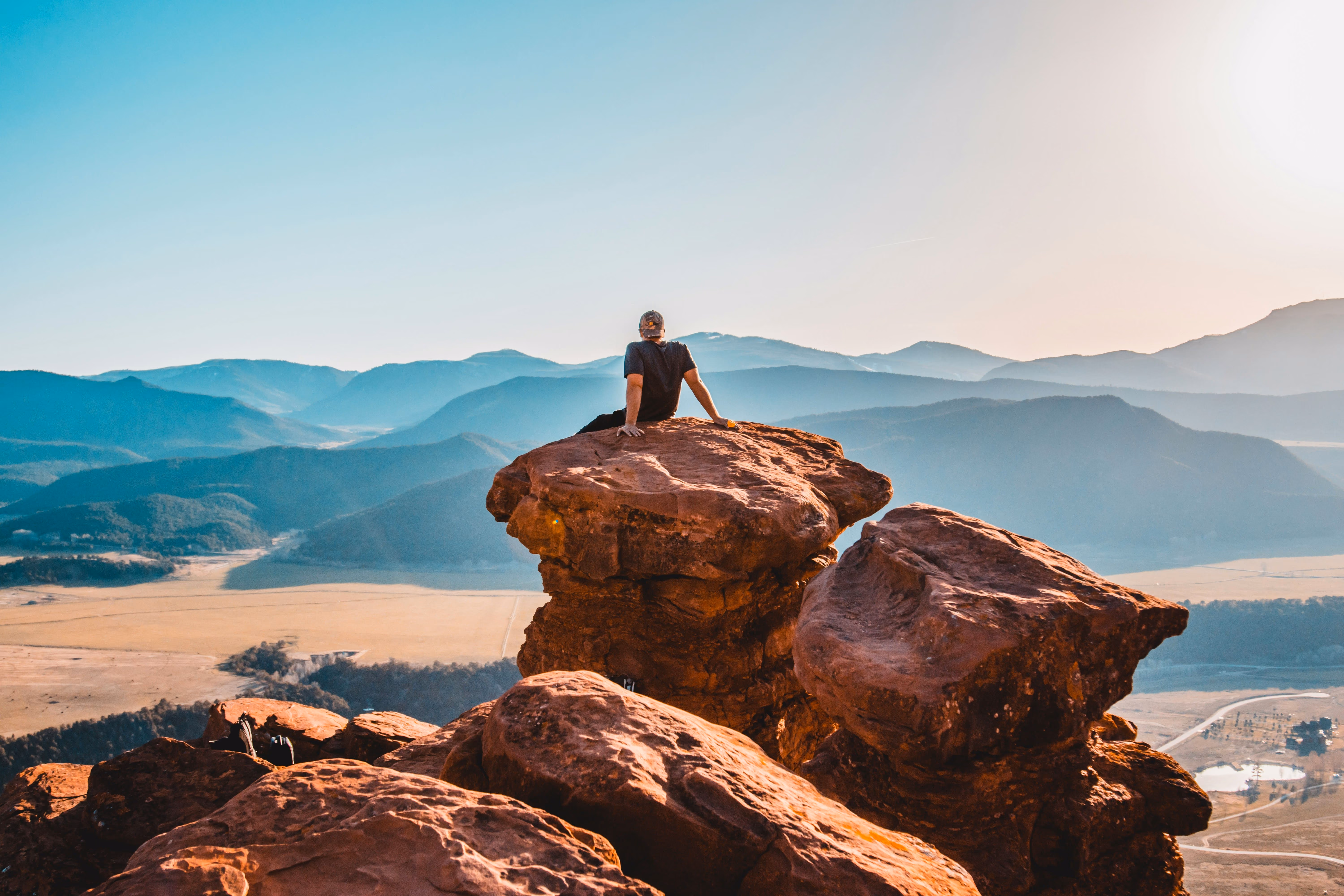 Person sitting on large rock overlooking a valley with distant mountain ranges at sunset.