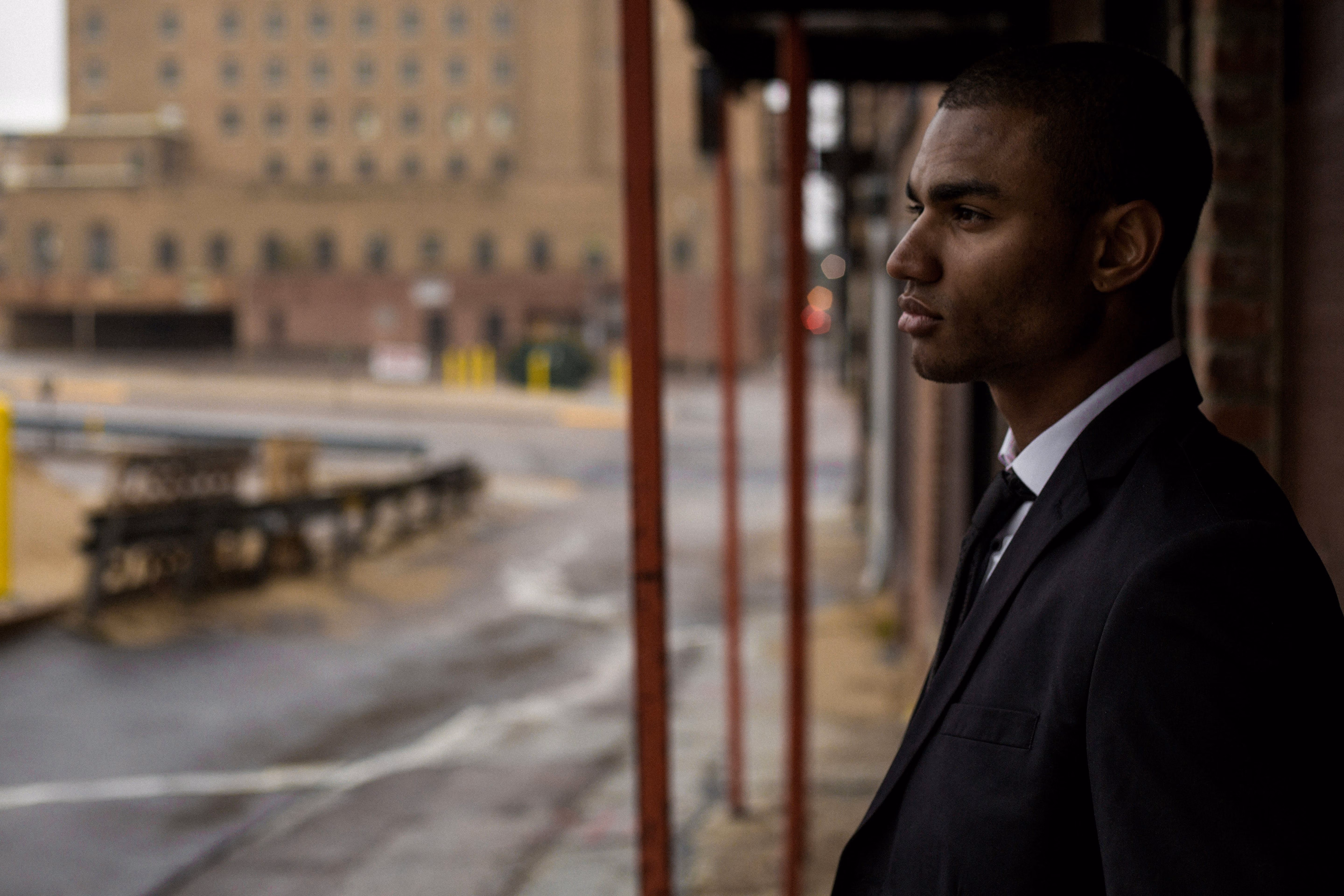 Young man in a black suit looking thoughtfully out at a blurred urban street scene.