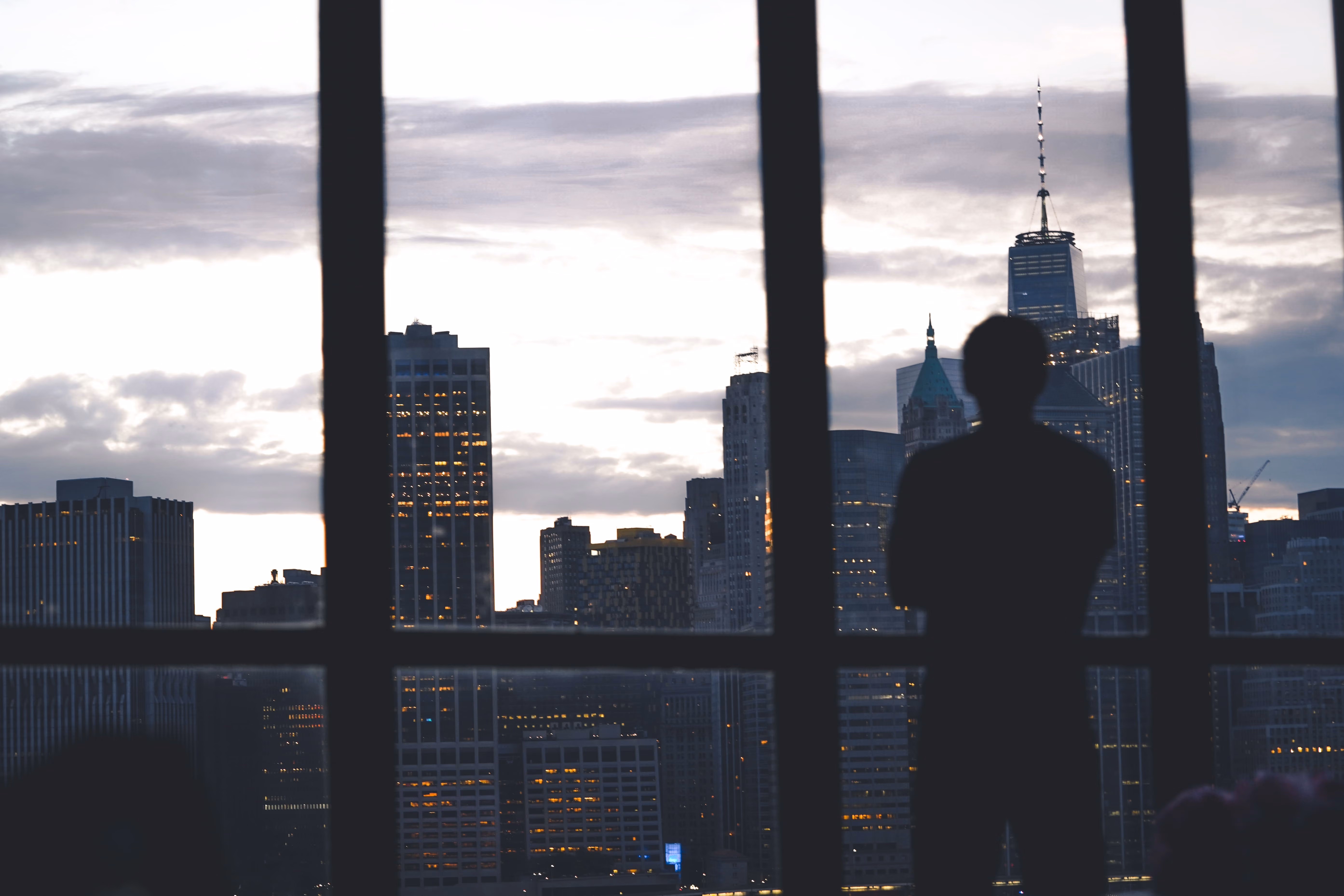 Silhouette of a man standing by a large window overlooking city skyscrapers at dusk.