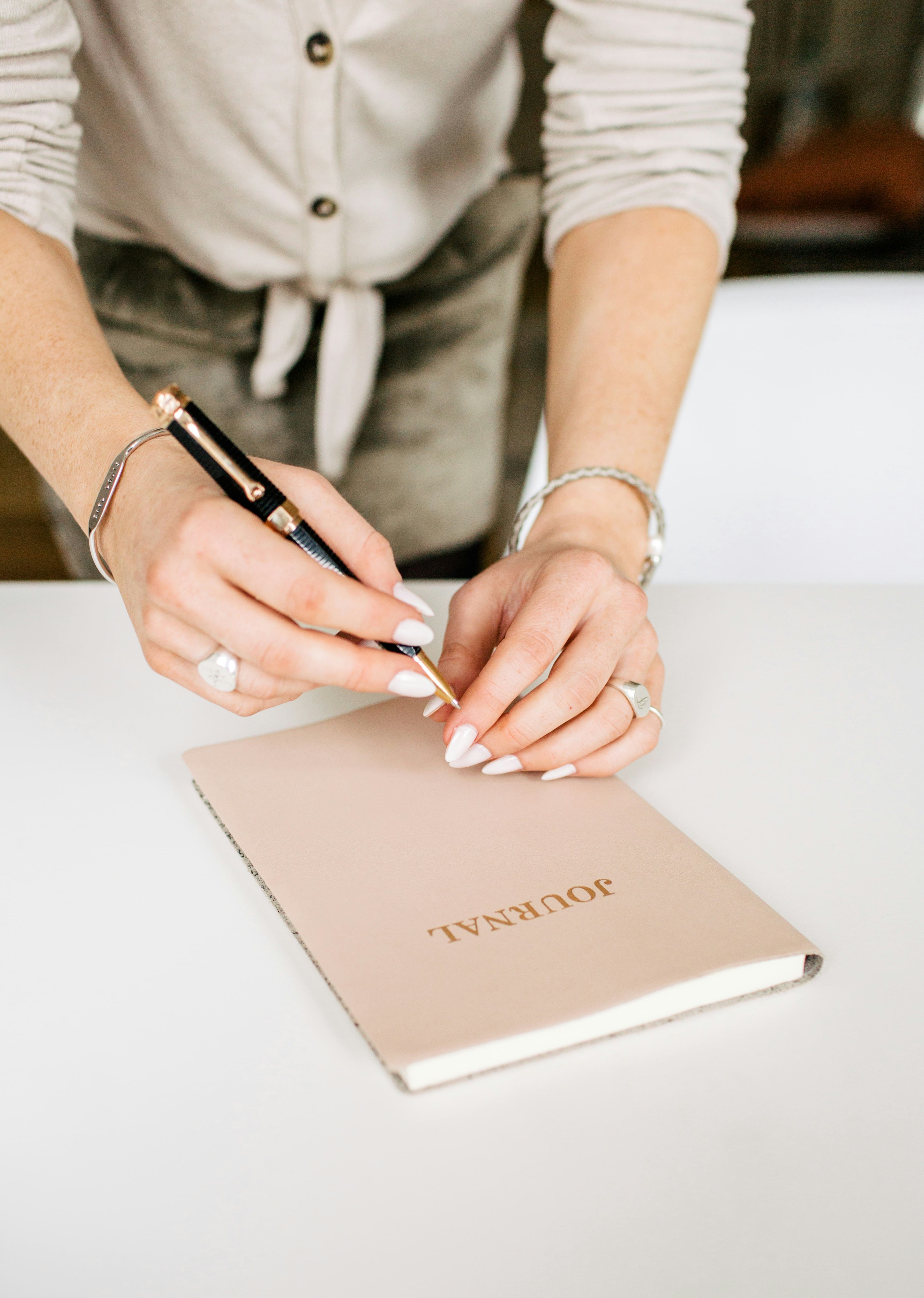 Person with manicured nails holding pen over a beige journal on a white table.