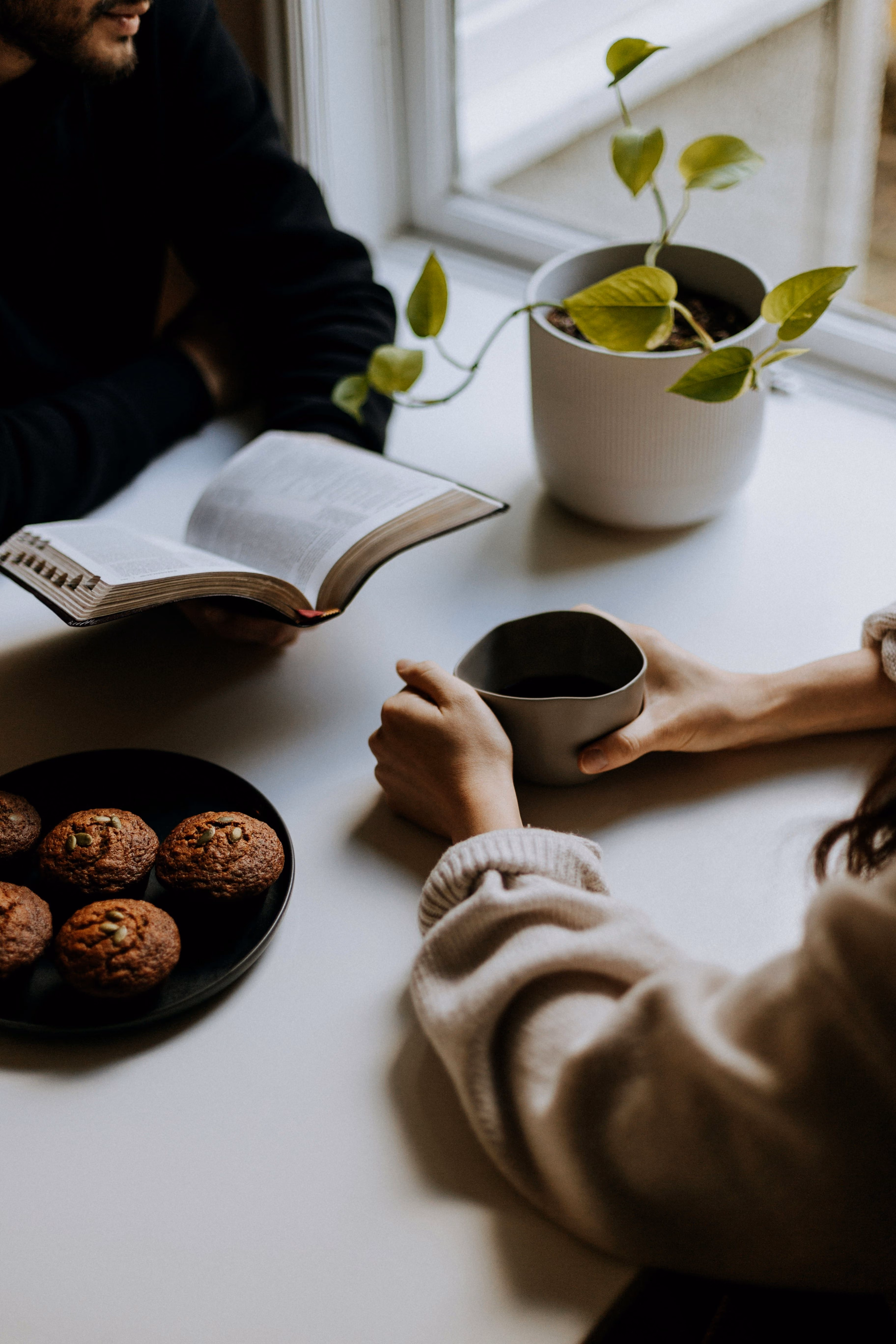 Two people at a table with one reading a book, the other holding a cup, muffins and plant nearby.