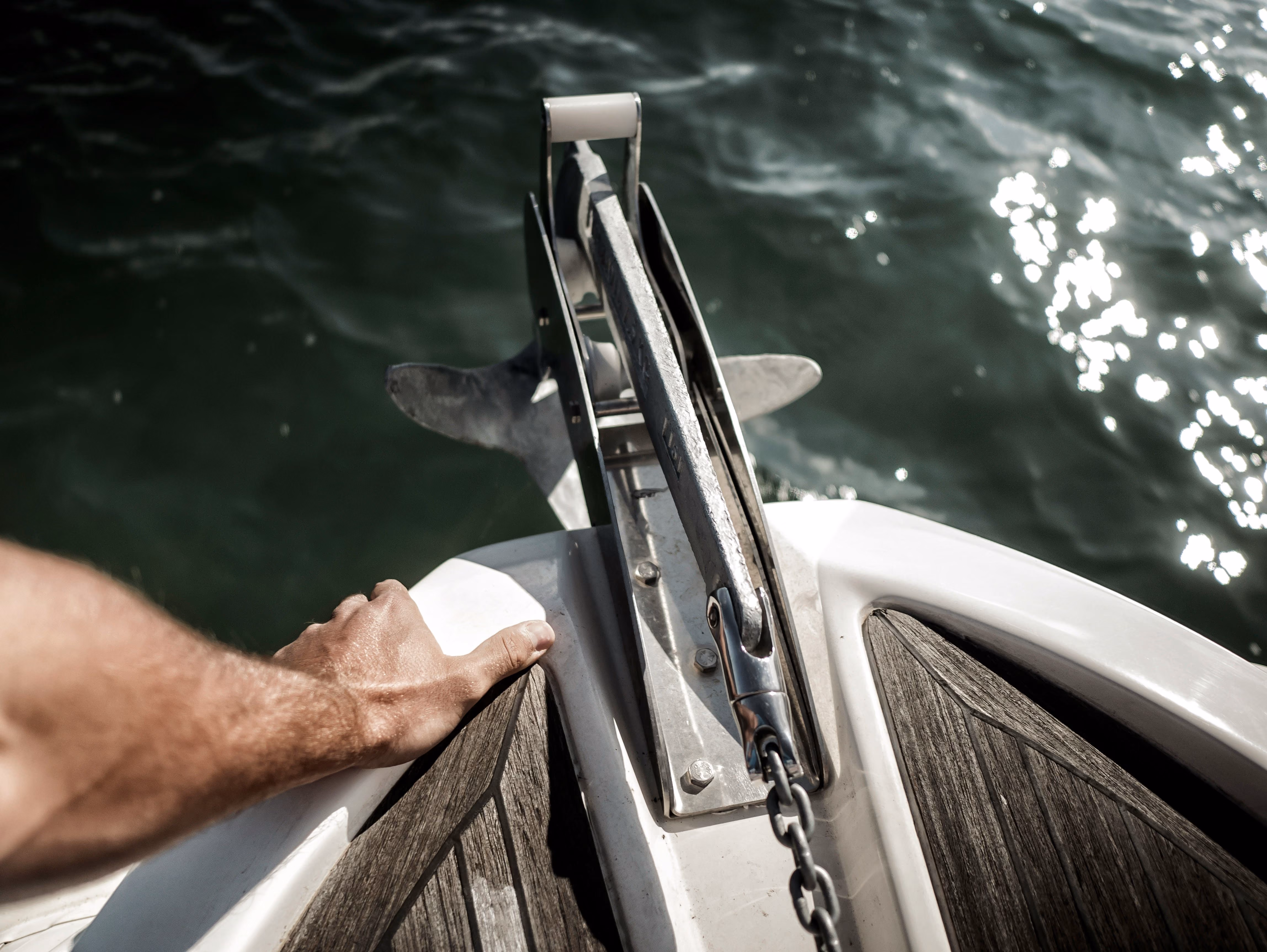 Hand resting on a boat deck near a metal anchor over dark, sparkling water.