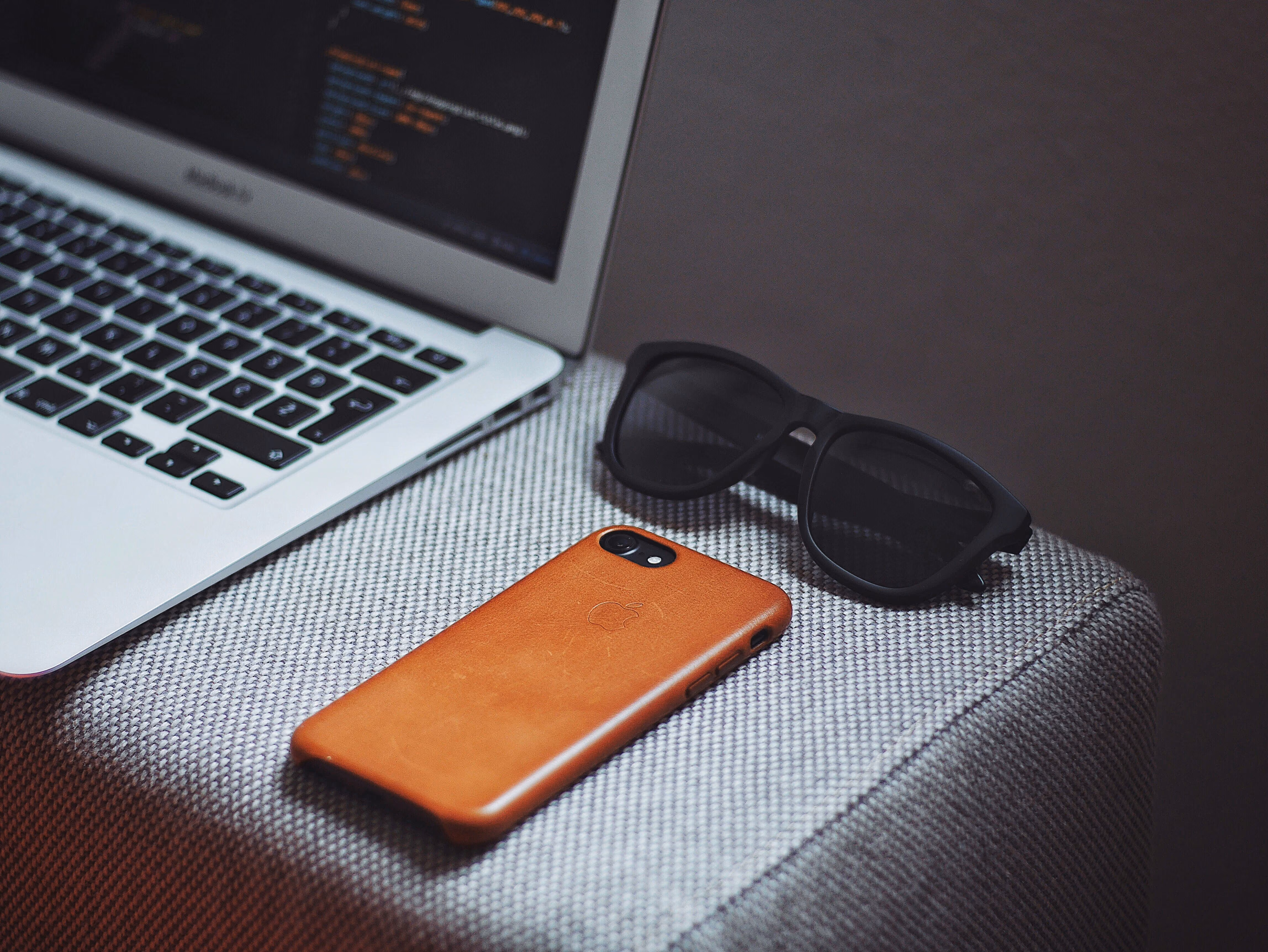 Laptop keyboard with coding on screen beside a phone with a brown case and black sunglasses on fabric surface.
