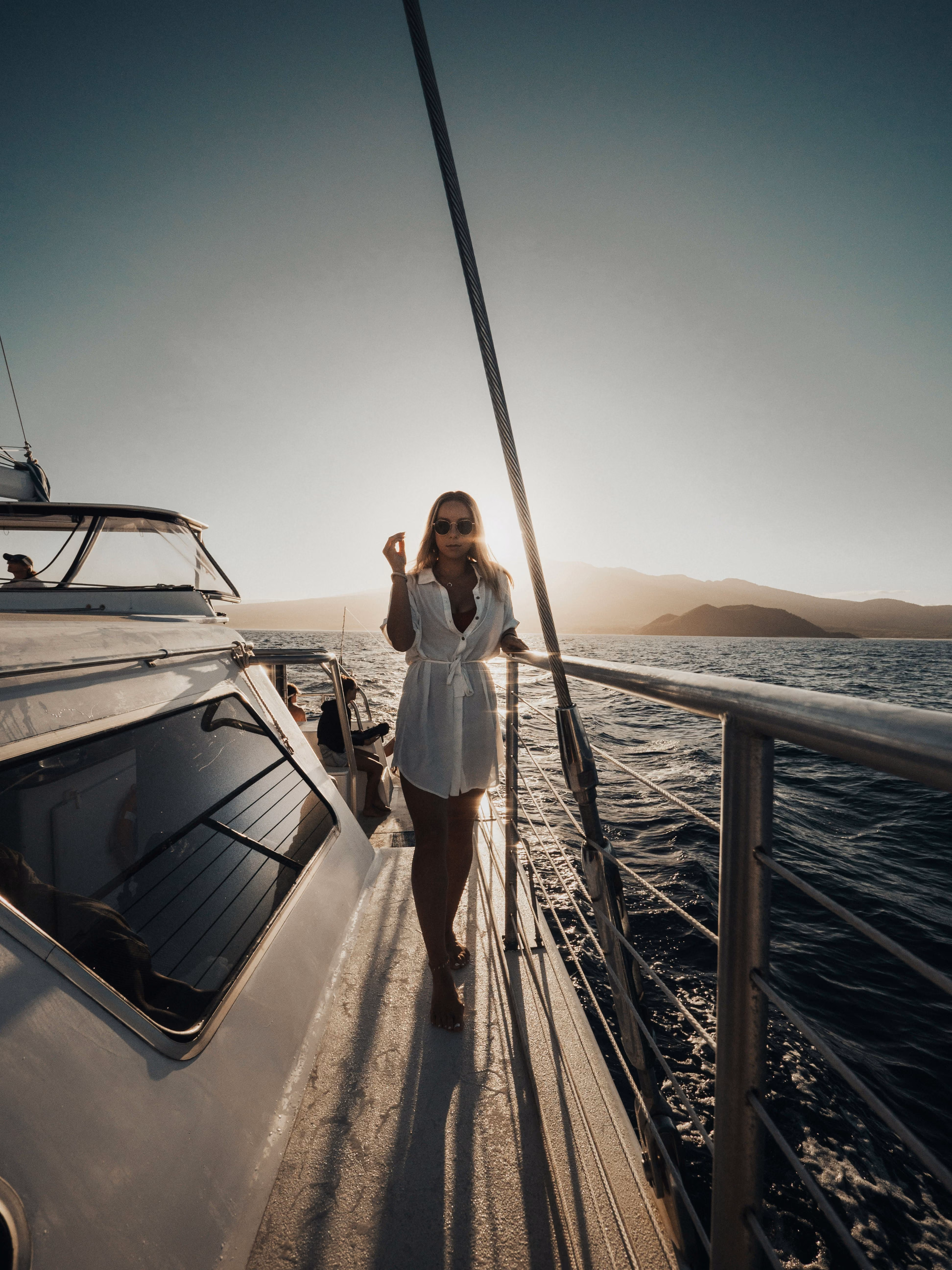Woman in white dress and sunglasses standing on a boat deck at sunset with mountains in background.