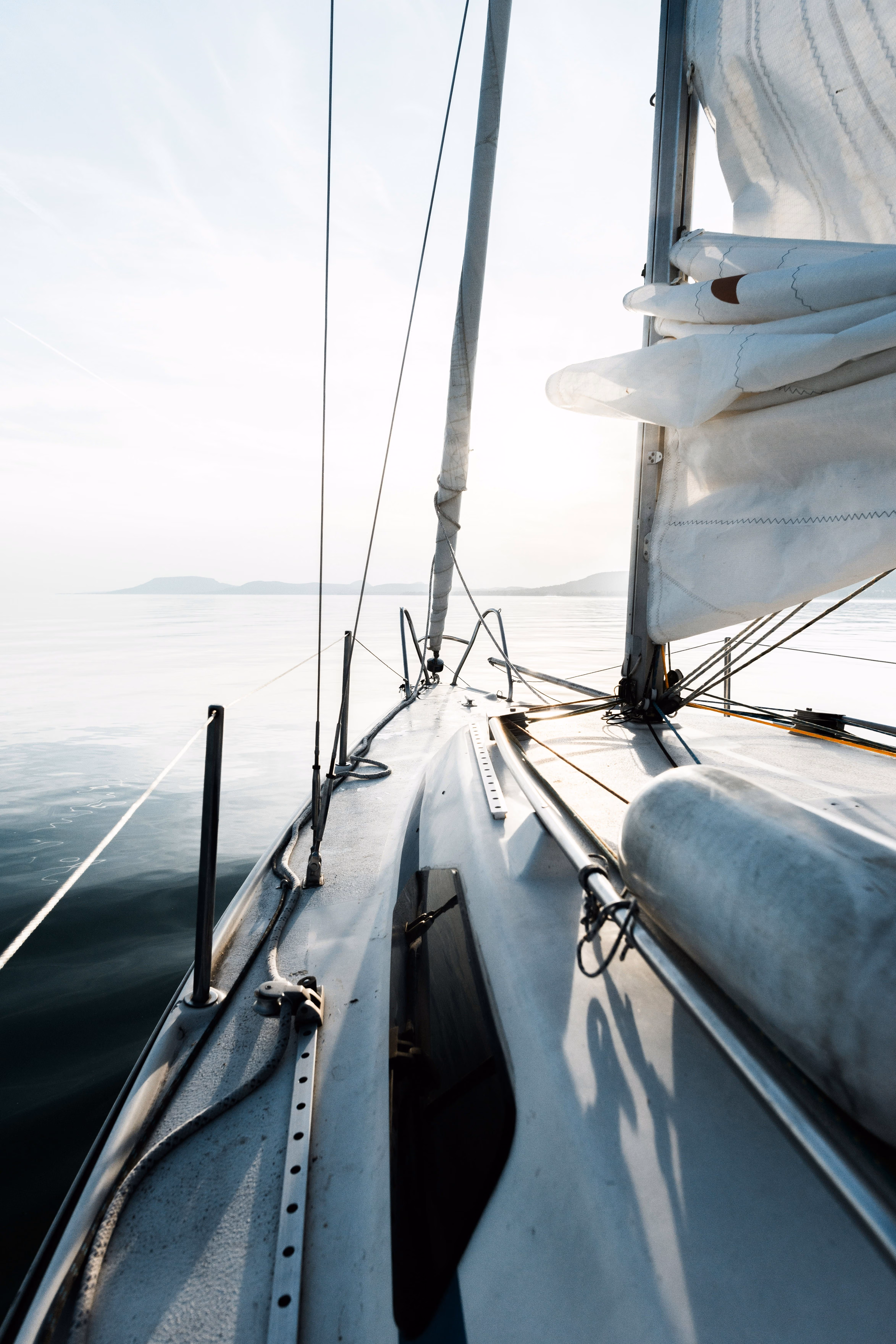 View from a sailboat deck showing rigging, sails, calm water, and distant mountains in soft light.