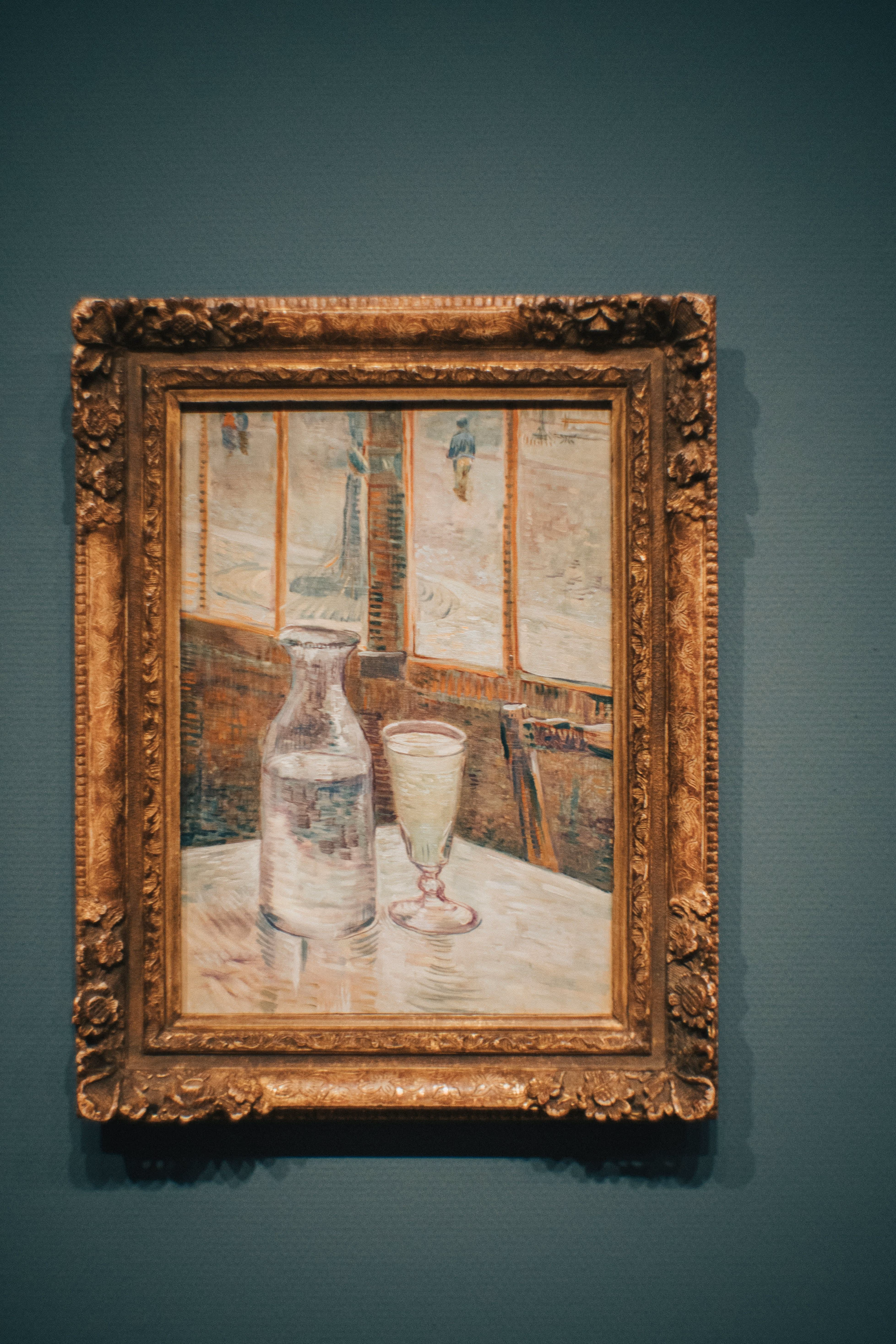 Painting showing a glass carafe and a goblet with water on a table by a window, in a decorative frame.