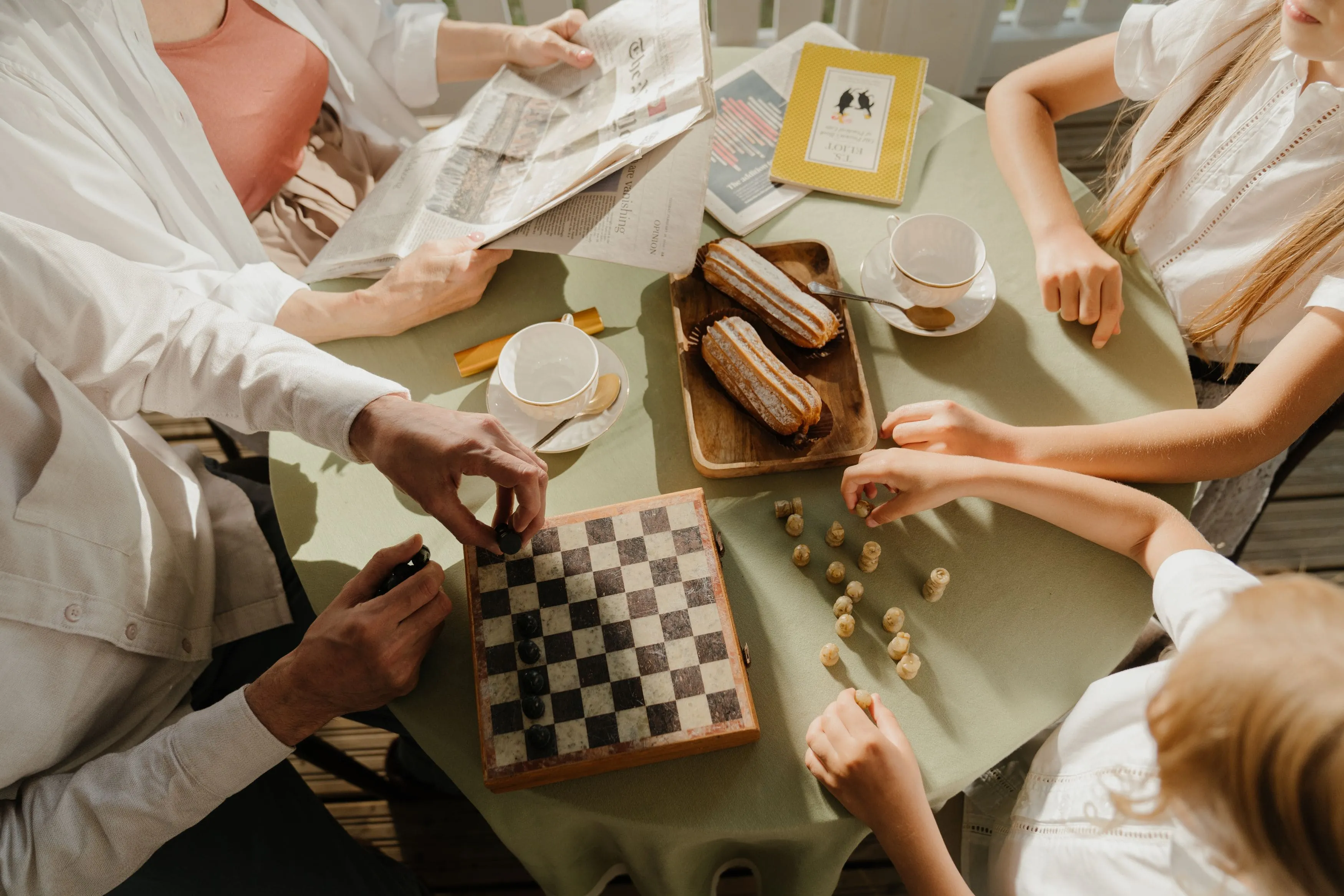 People playing chess at a table with tea cups, books, and two eclairs on a wooden tray.