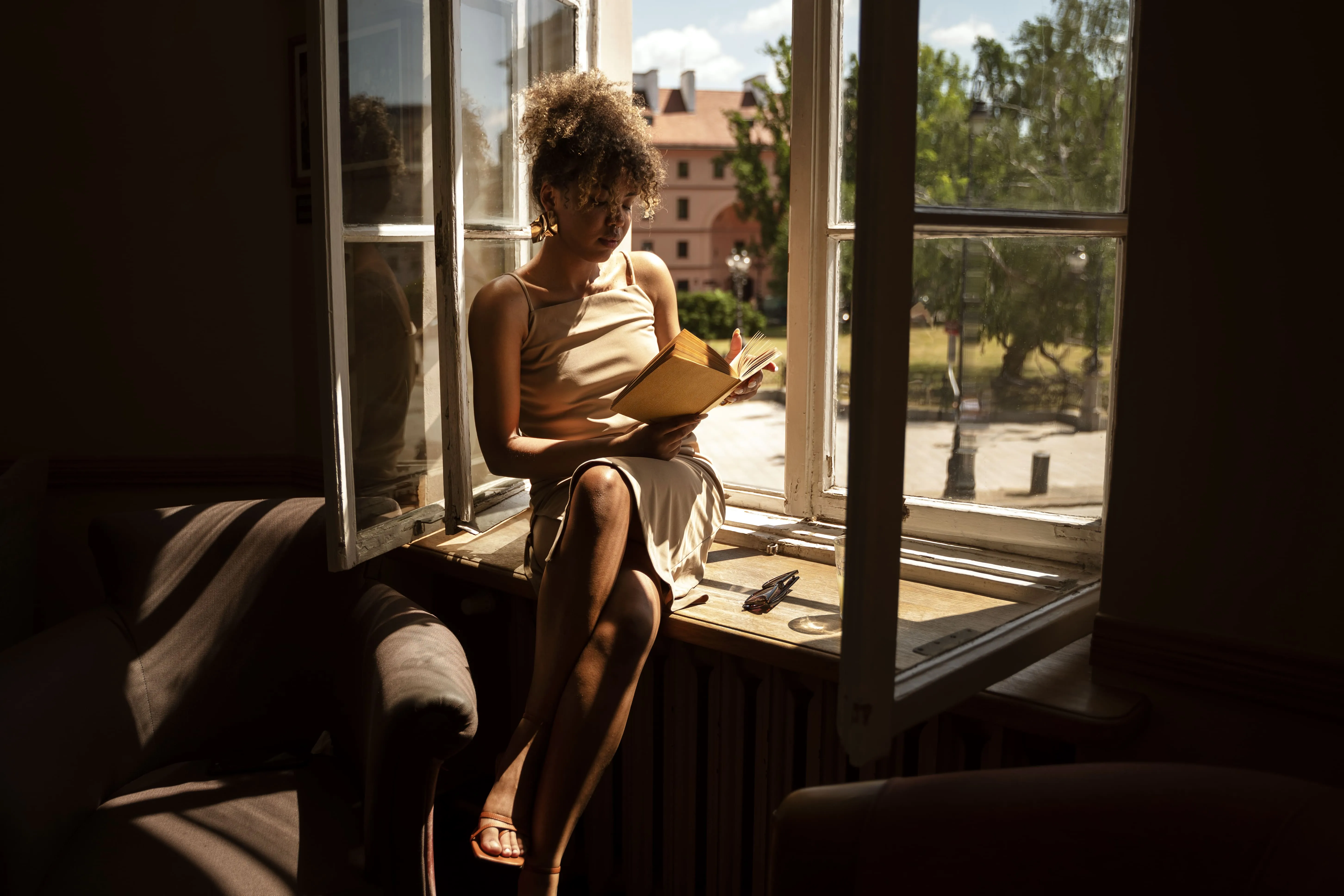 Woman with curly hair reading a book sitting on a windowsill with sunlight outside.
