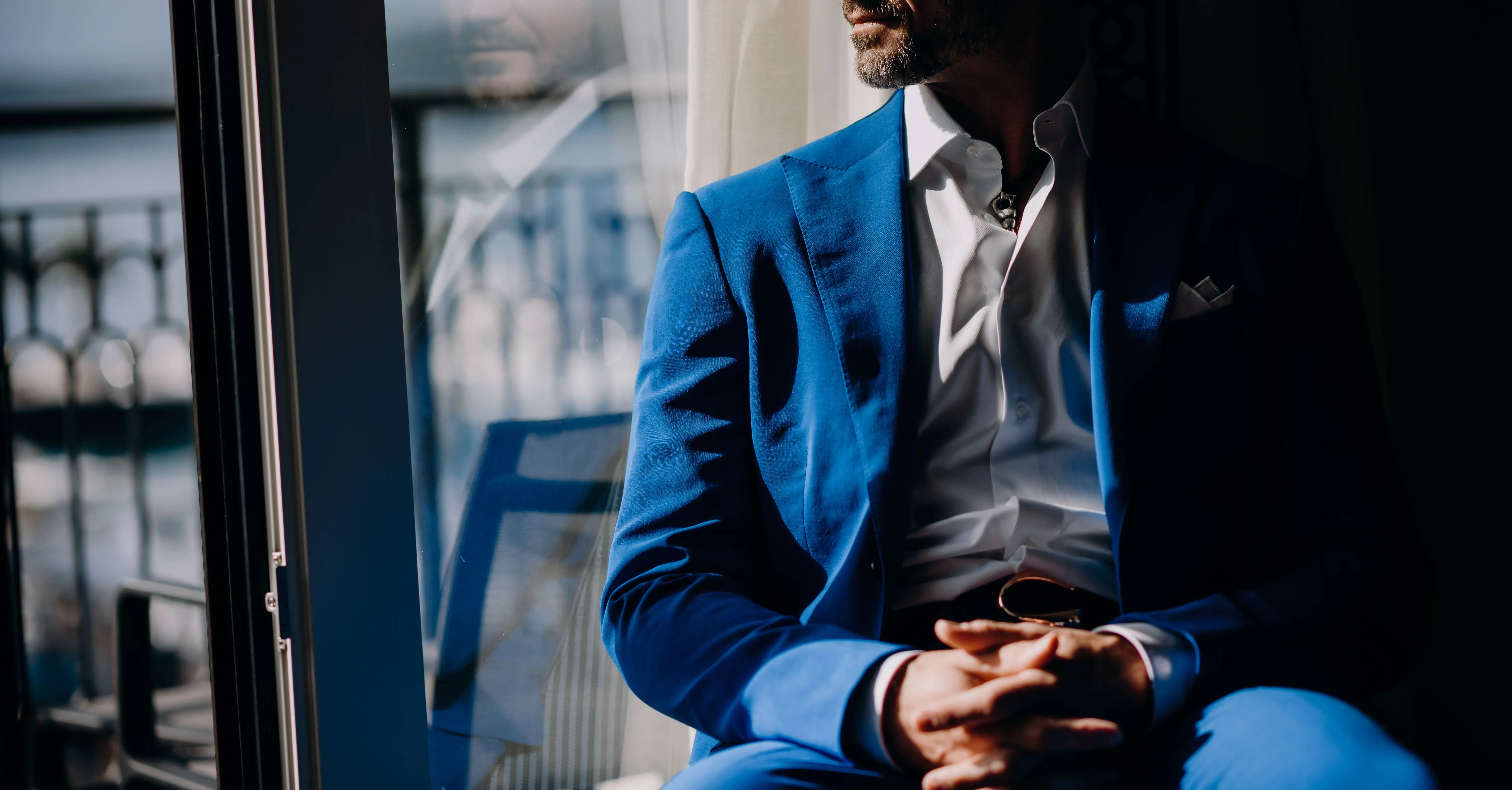 Man in blue suit sitting by a window with hands clasped and partially visible face looking outside.