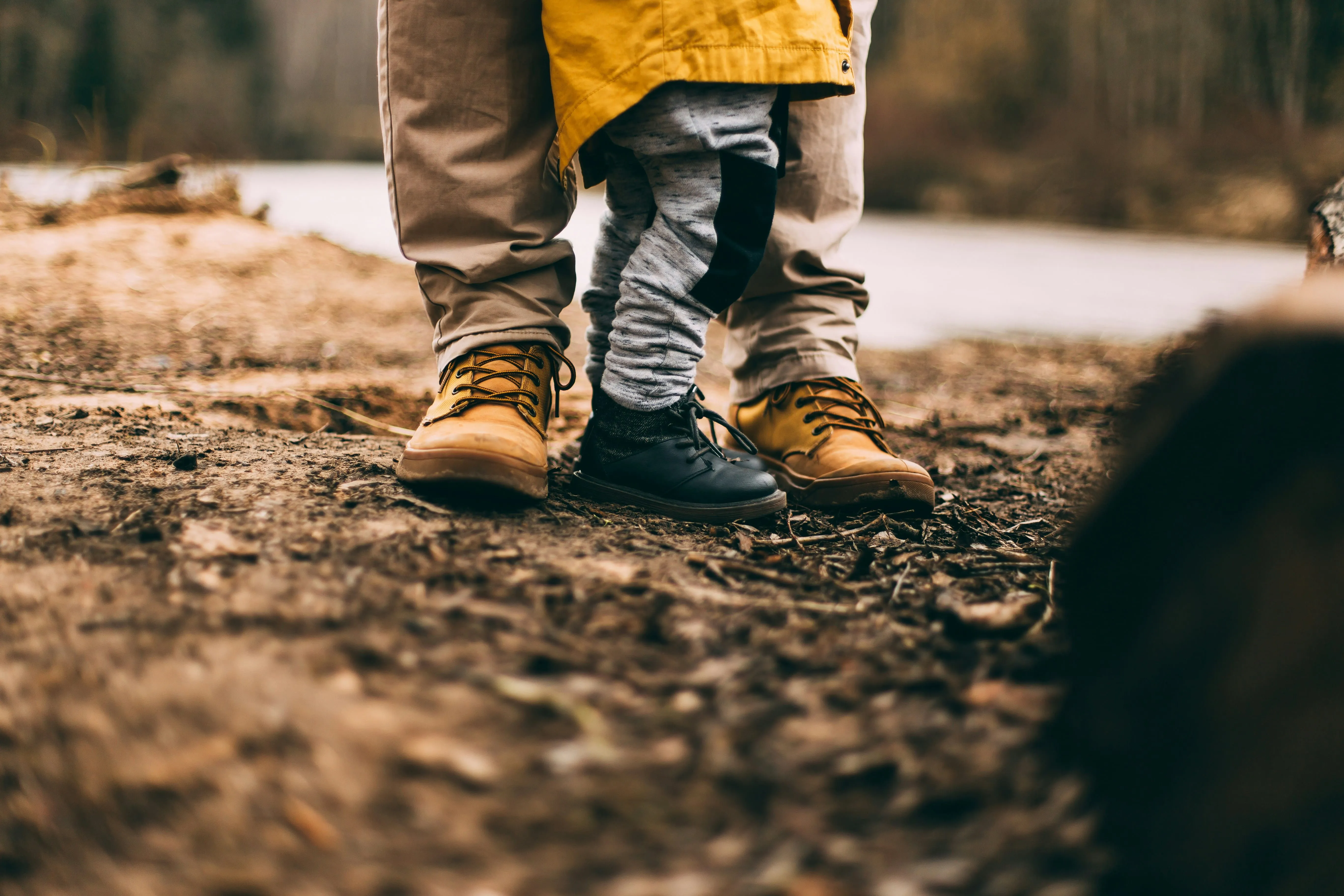 Adult wearing tan boots stands behind child in black shoes and gray pants on dirt ground outdoors.