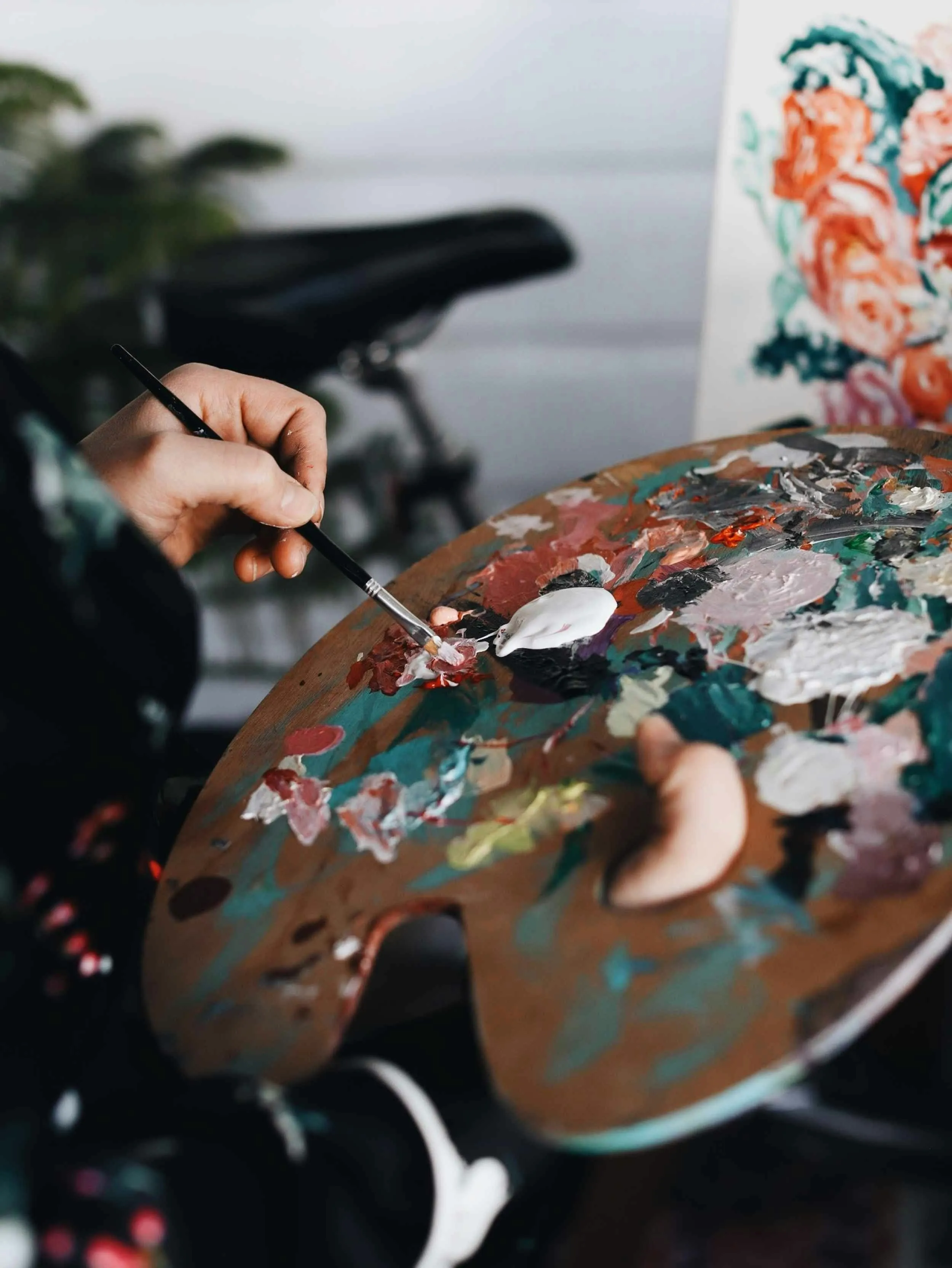 Hand holding a brush mixing paint on a colorful artist's palette with a floral painting in the background.
