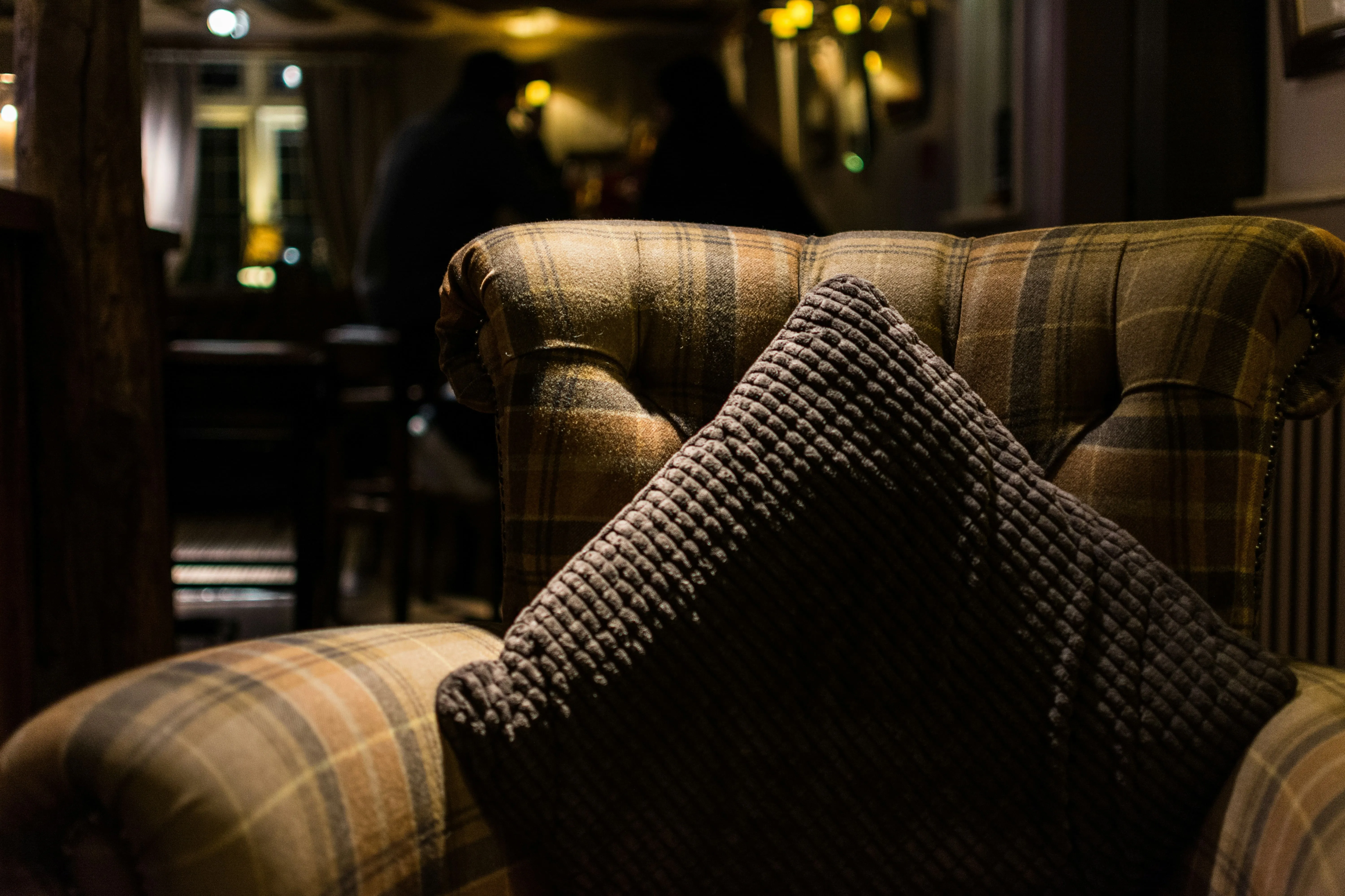 Plaid armchair with a dark textured cushion in a dimly lit cozy room with blurry background figures.