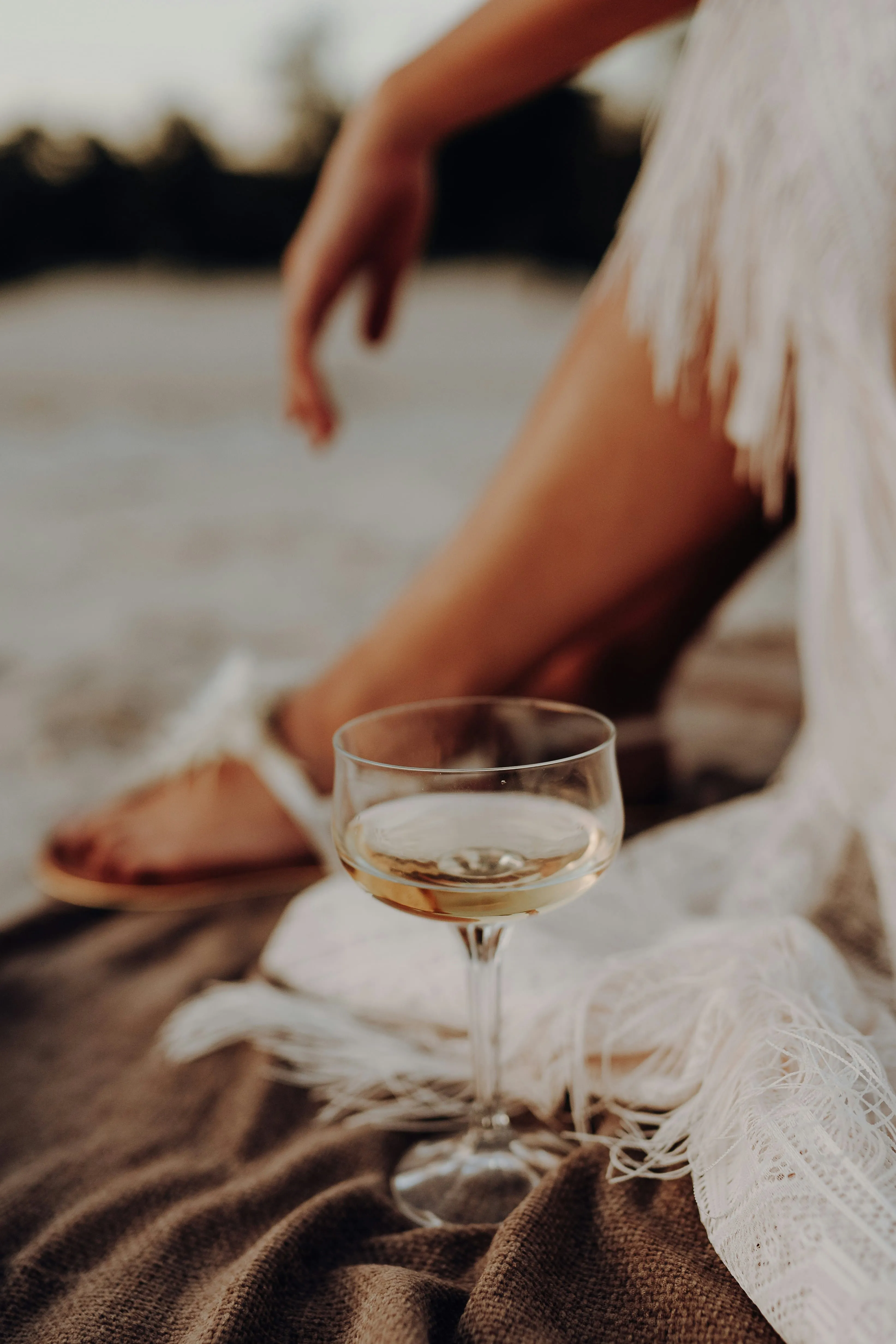 Close-up of a glass of white wine on a blanket near a person wearing a white dress and sandals.