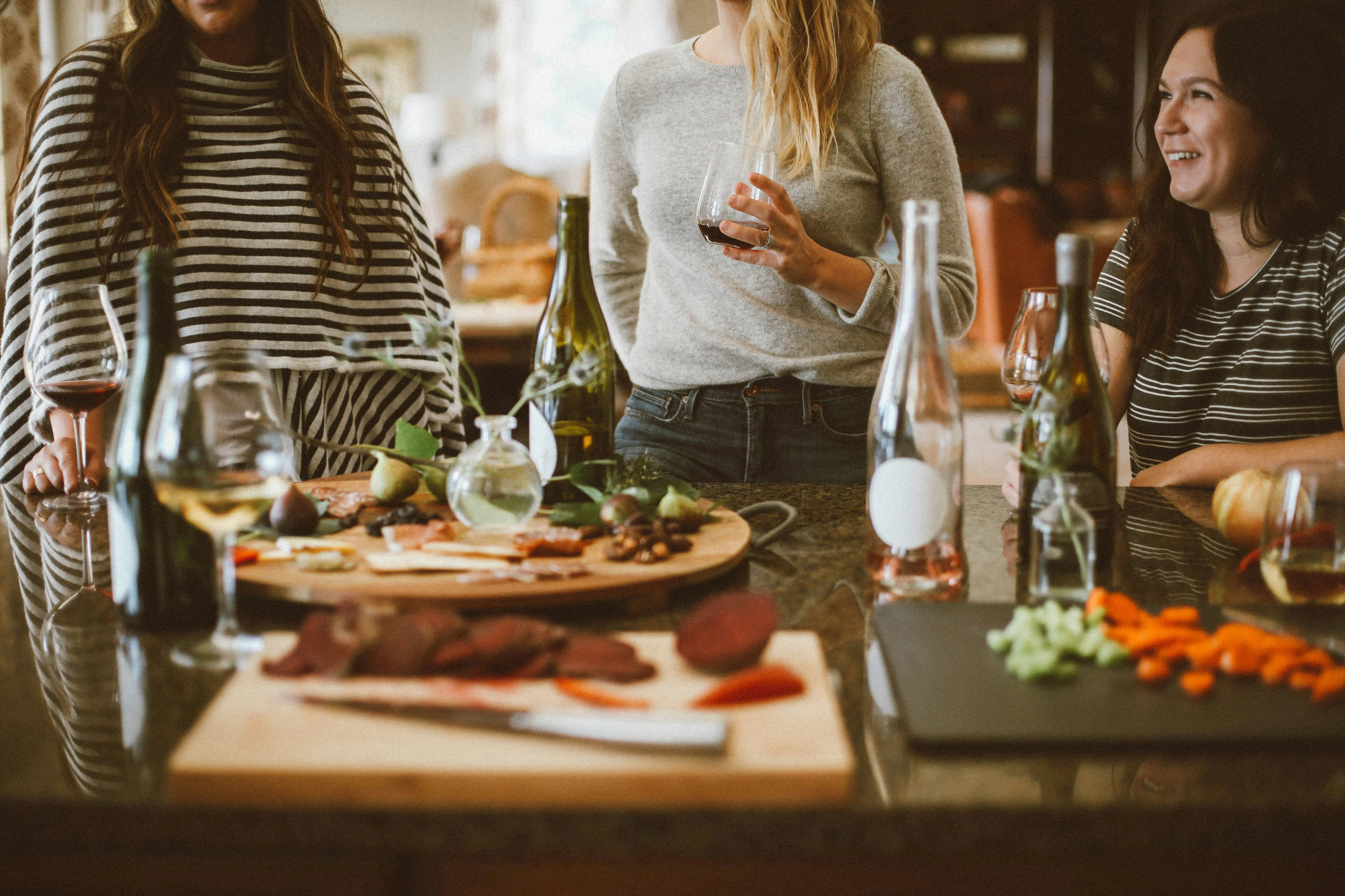 Three women standing by a kitchen counter with wine glasses, bottles, and snacks on cutting boards.