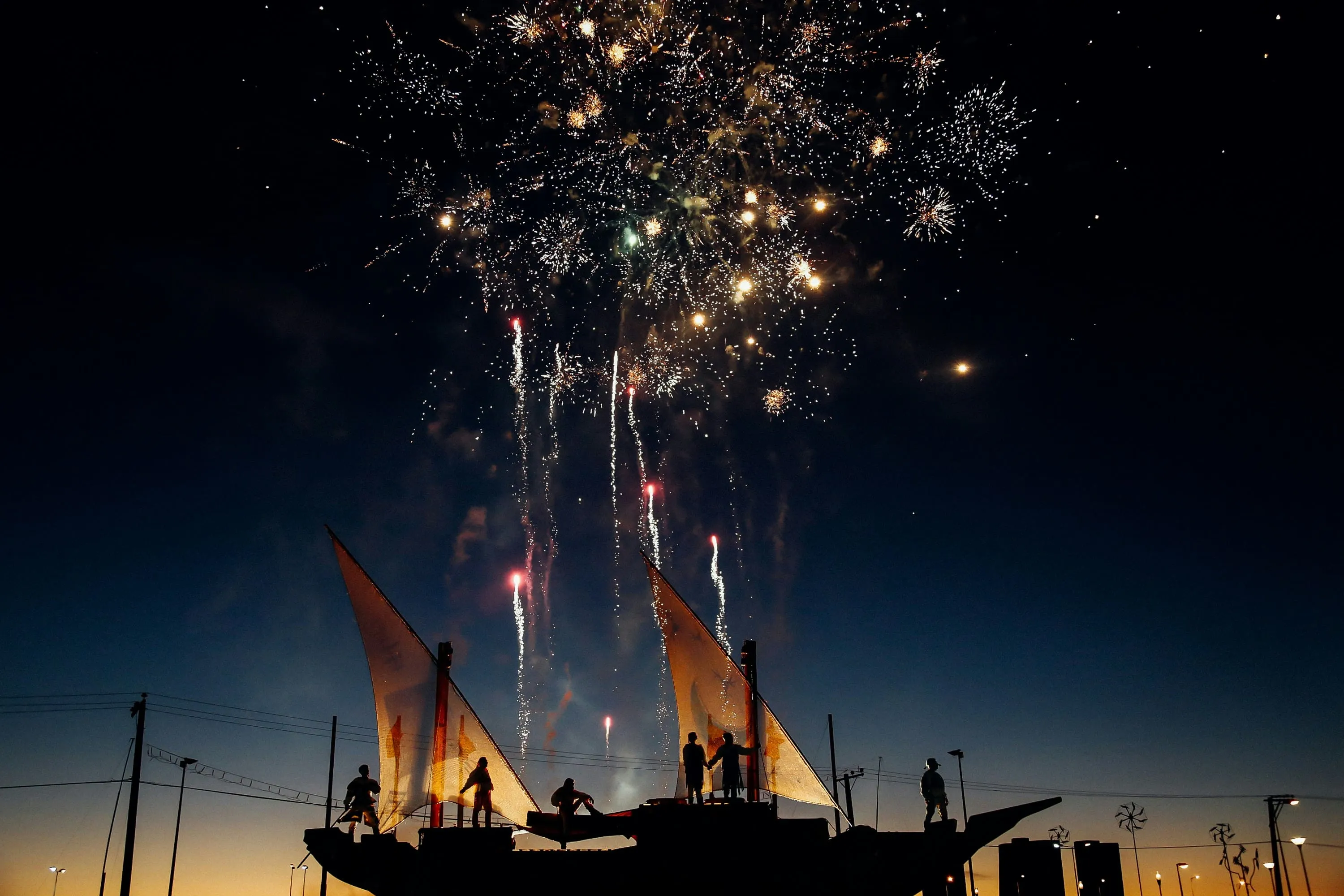 Silhouettes on a sailboat watch fireworks burst in the night sky at dusk.