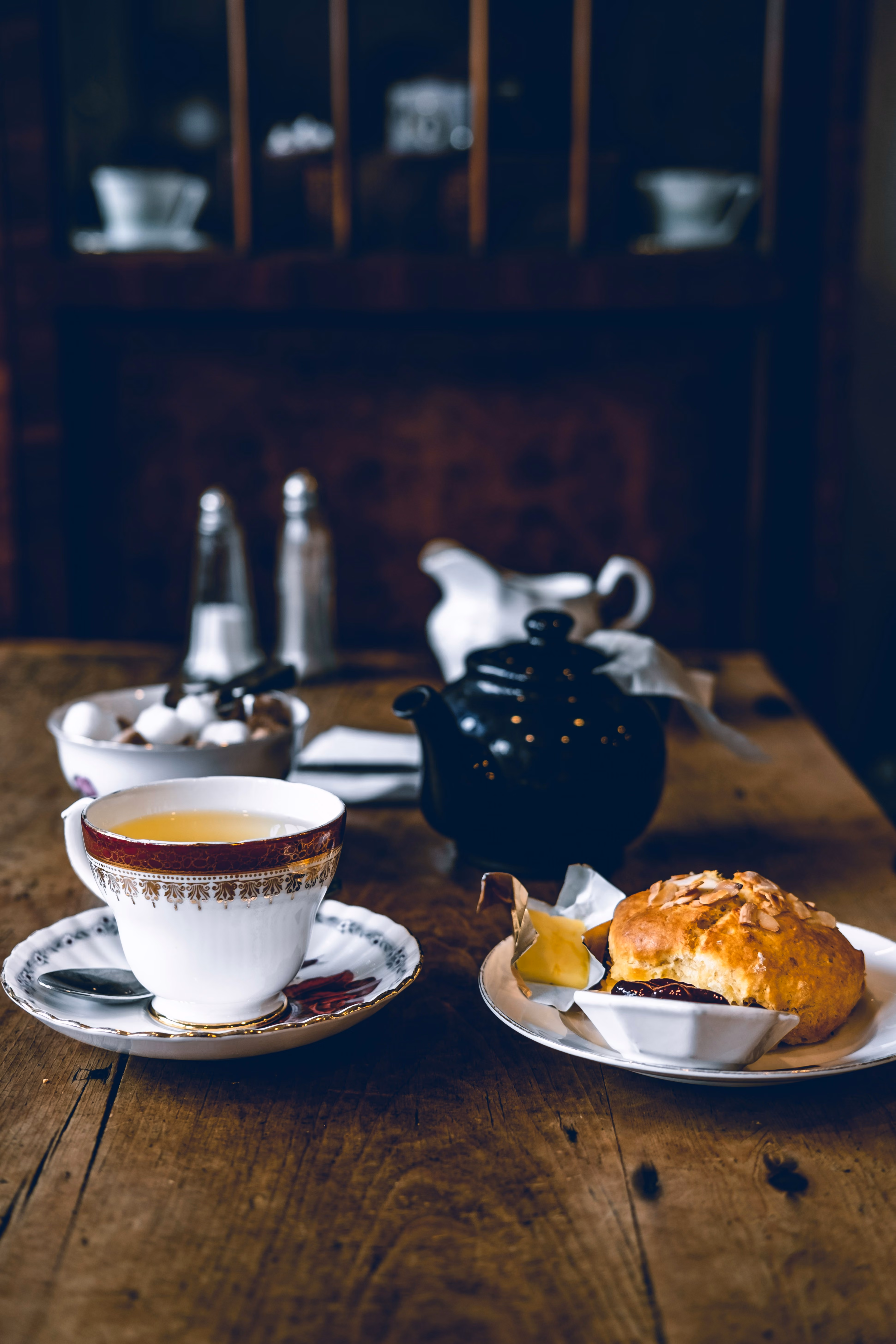 Tea cup with saucer, scone with jam and butter, black teapot on wooden table.