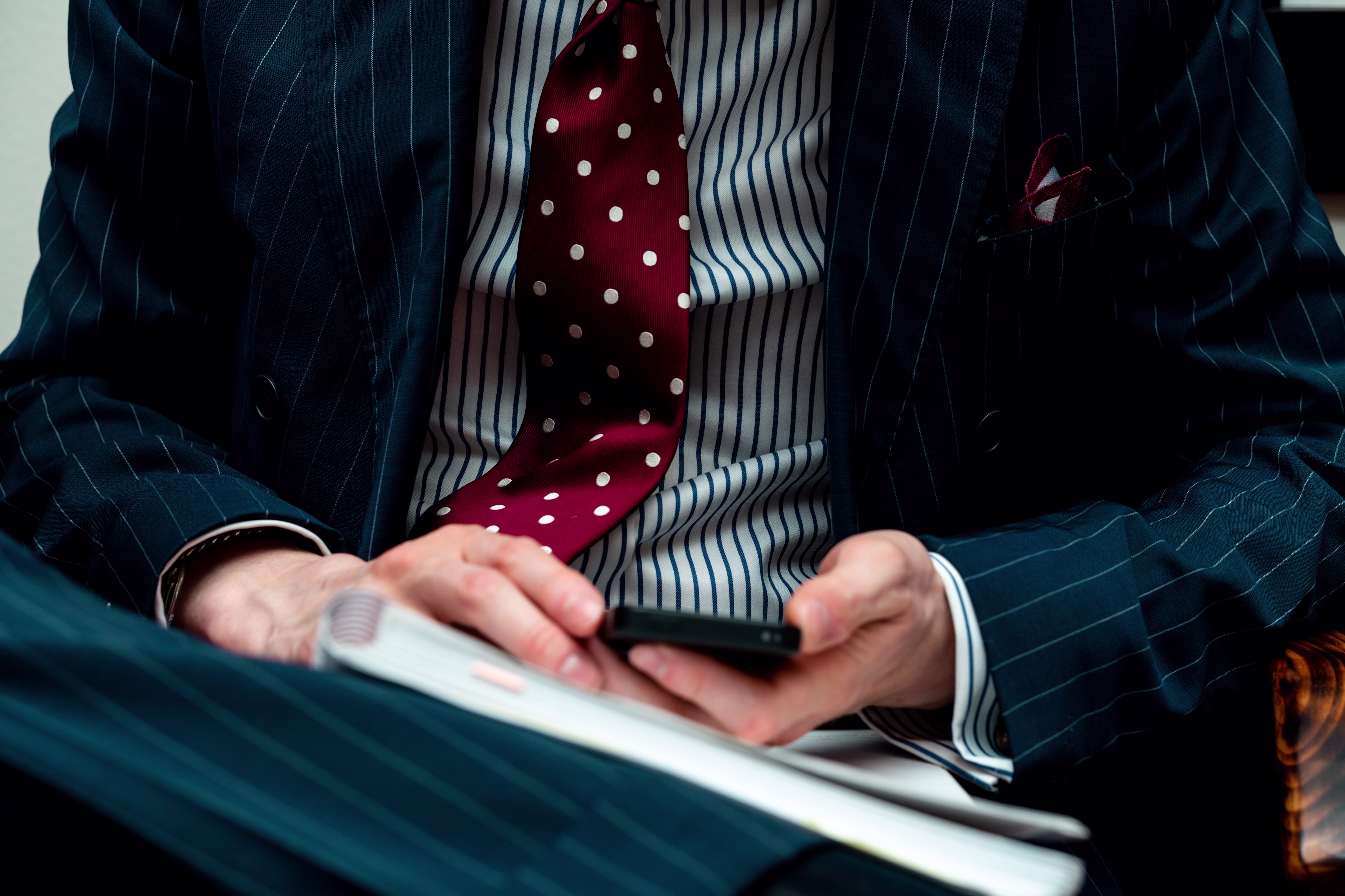Person in pinstripe suit and red polka dot tie holding a phone and notebook.