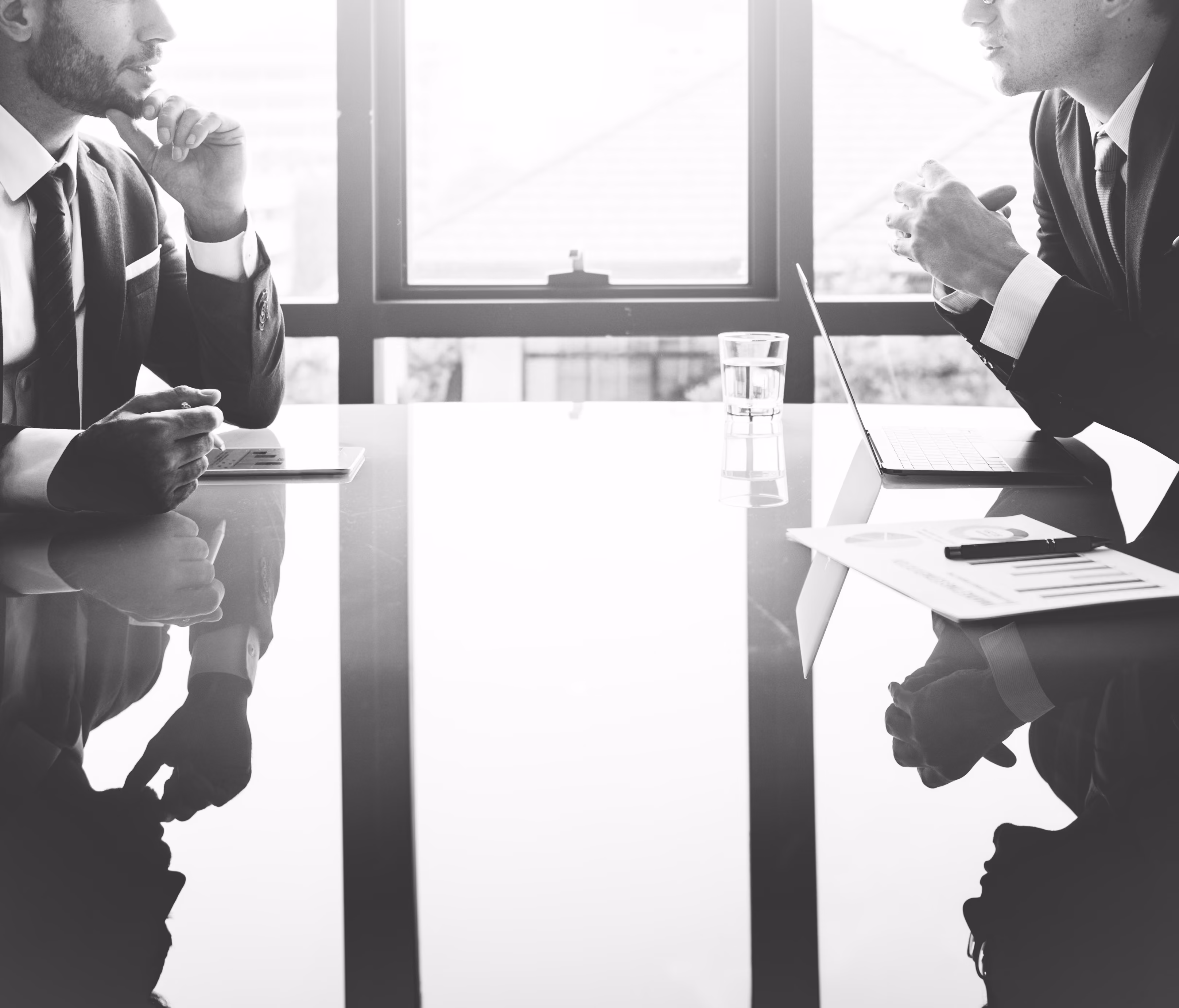 Two businessmen in suits sitting at a reflective table with documents and a laptop, talking.