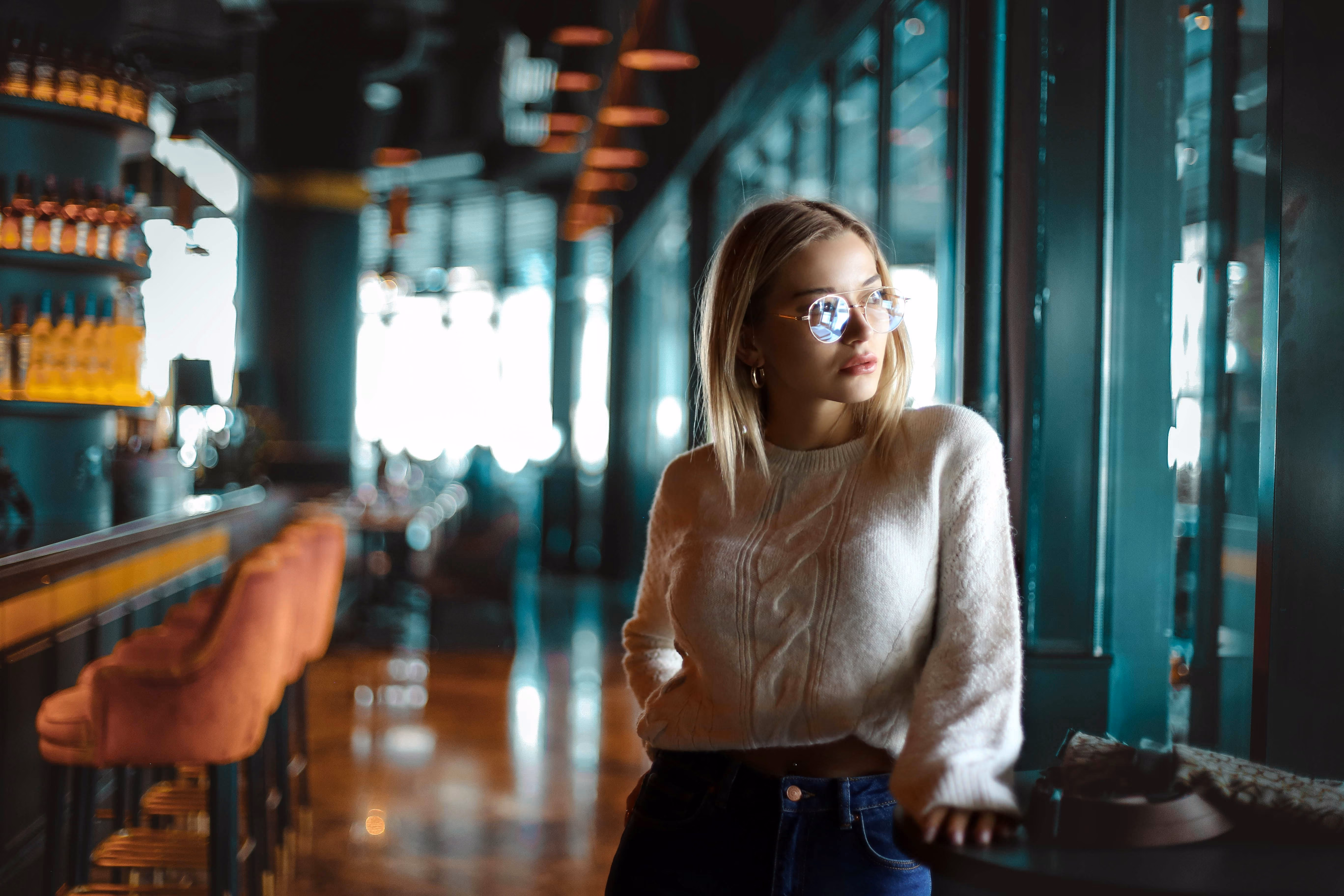 Woman in glasses and white sweater standing by a window in a dimly lit bar with pink stools.