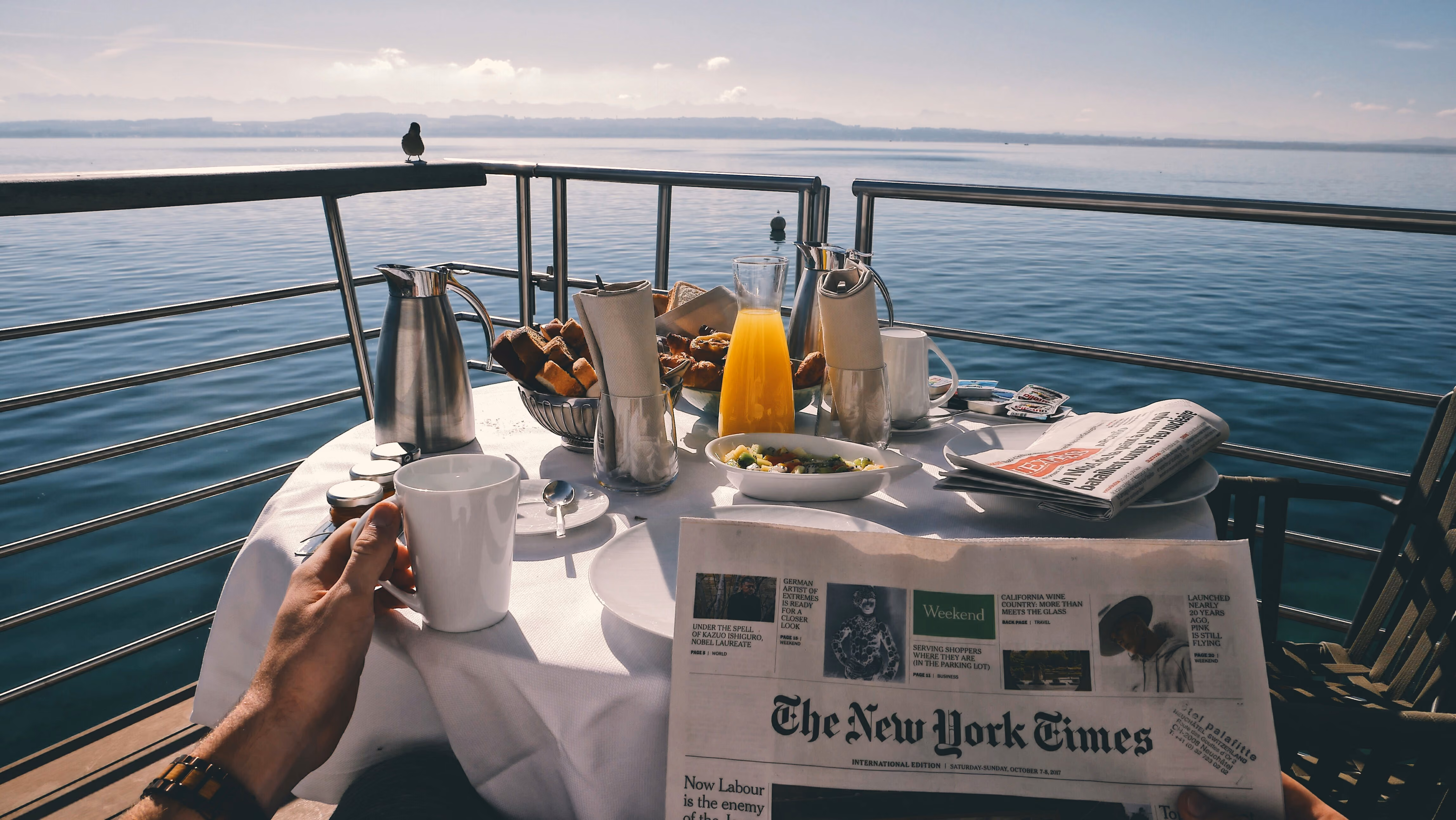 Person holding a coffee cup and reading The New York Times at a breakfast table by the water.