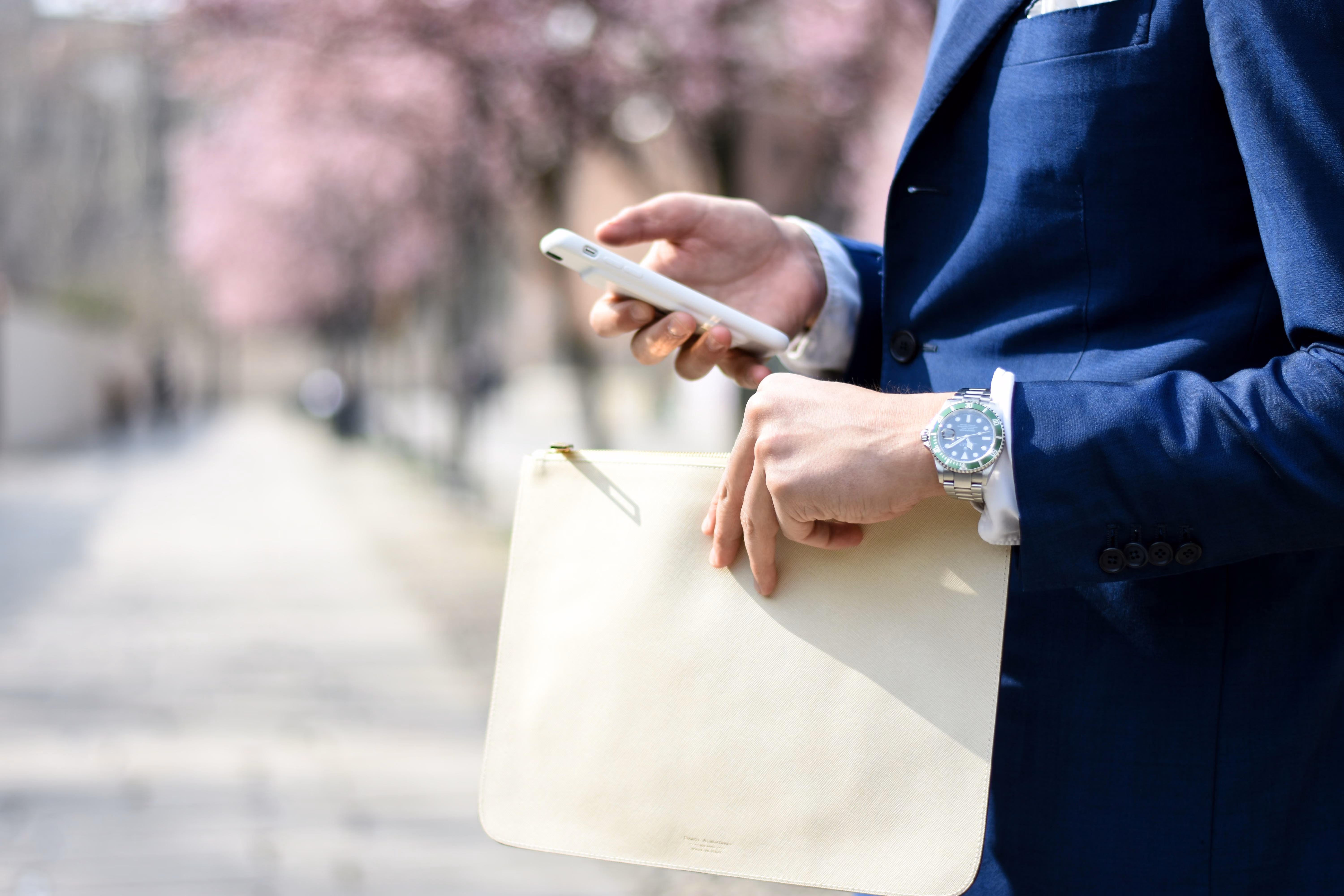 Person in navy suit holding a white phone and beige clutch outdoors with blurred pink trees.