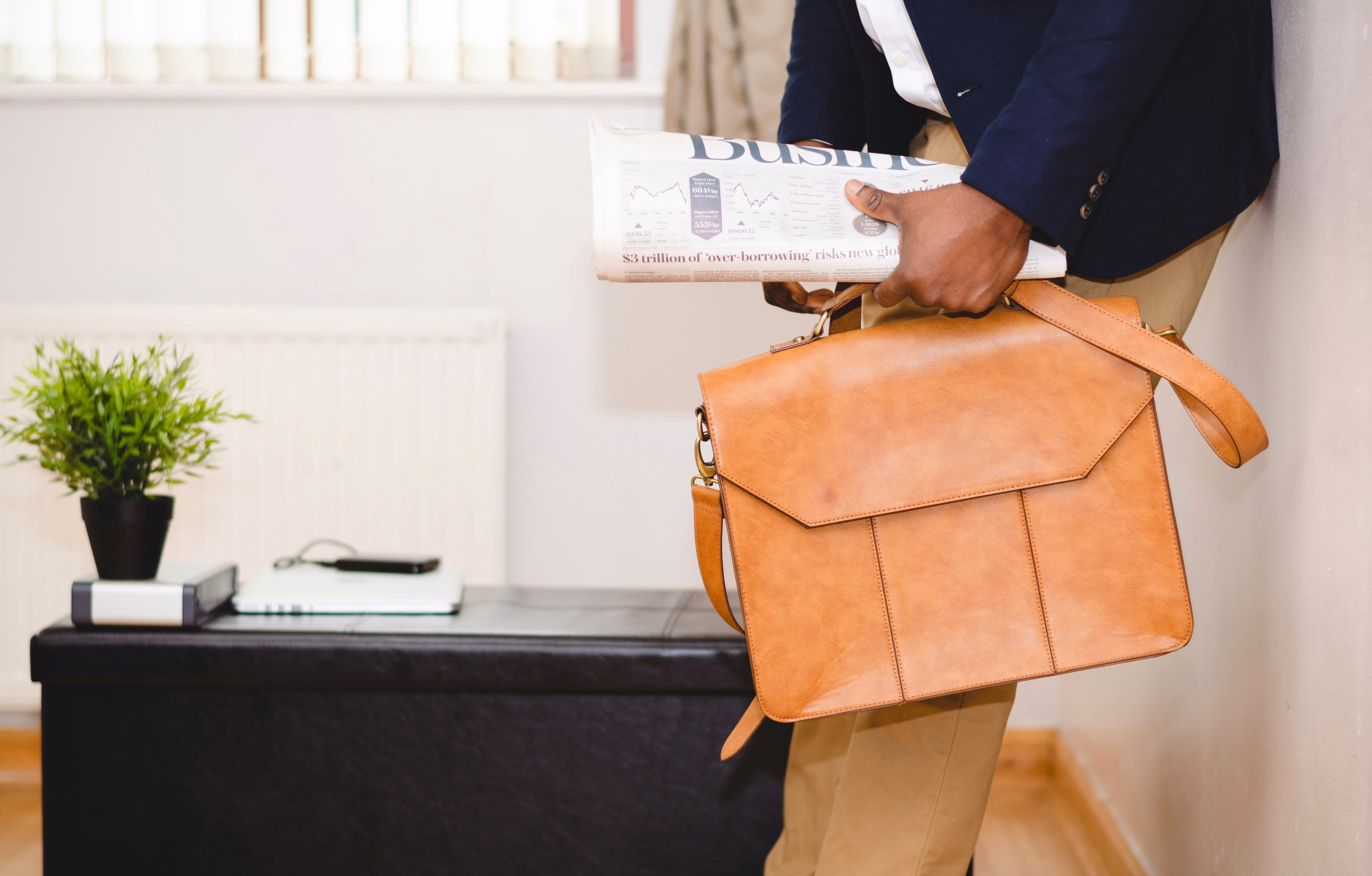 Person holding a rolled newspaper and a tan leather briefcase, standing indoors.
