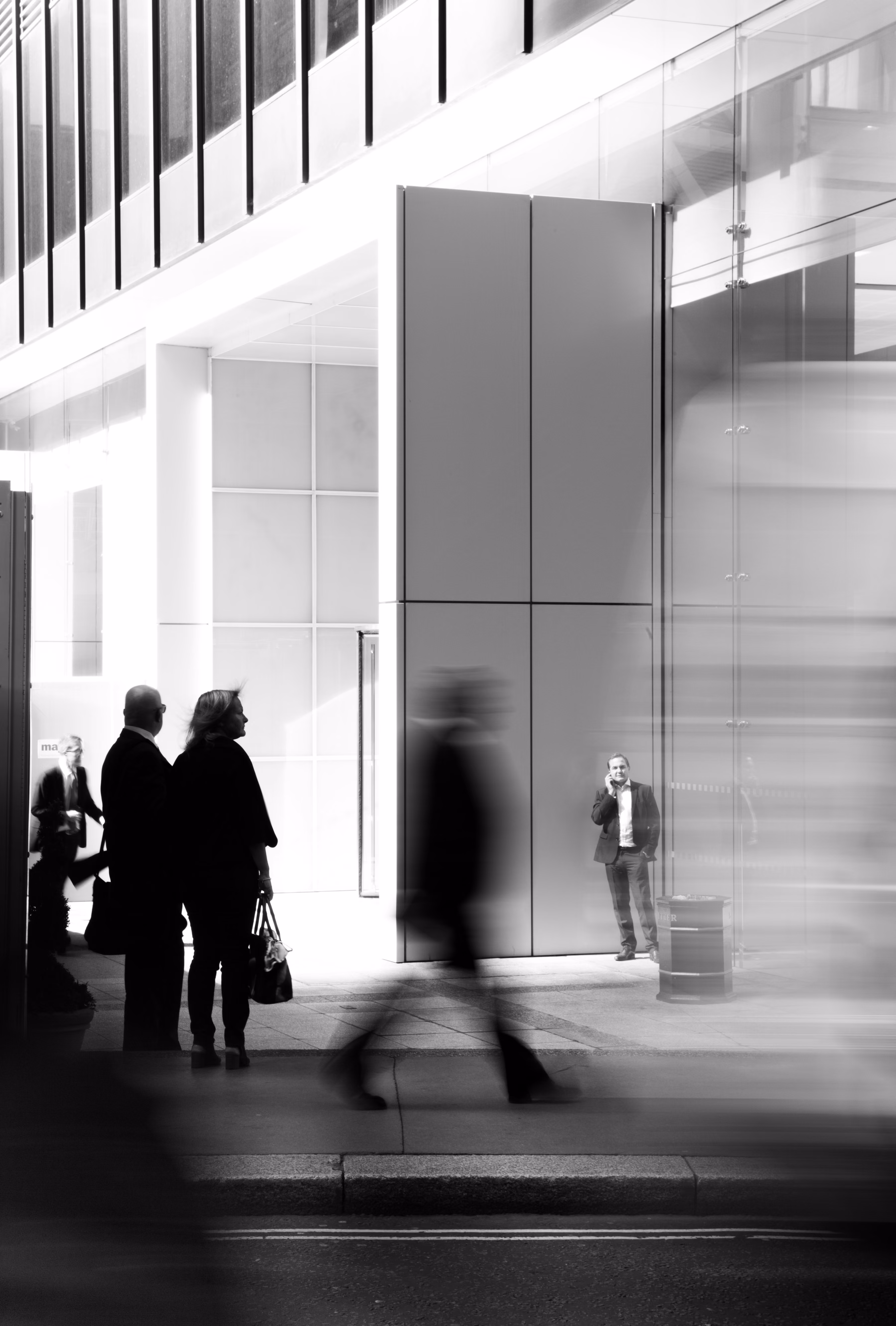 Black and white photo of people walking and standing near a modern building with glass and panels.