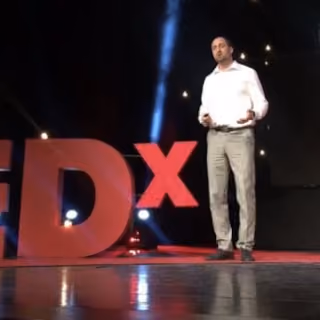 Man in white shirt and gray pants speaking on stage next to large red TEDx letters.