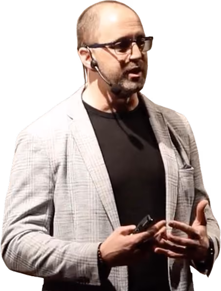 Man with glasses and headset speaking, wearing a gray plaid blazer and black shirt.
