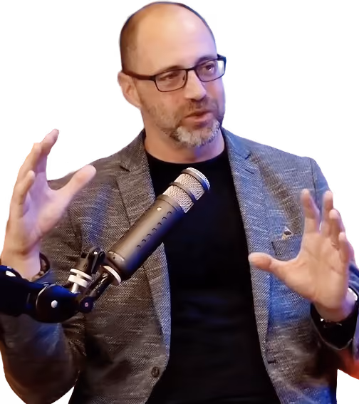Man with glasses and beard speaking into microphone, gesturing with both hands.