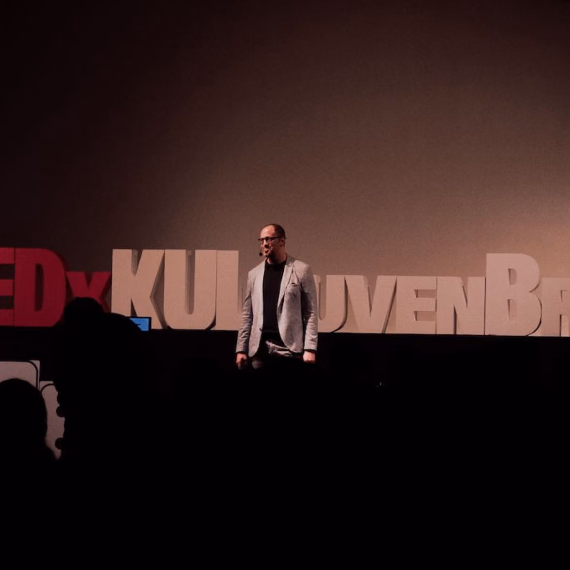 Man in light blazer speaks on stage at TEDx event with large letters behind him.